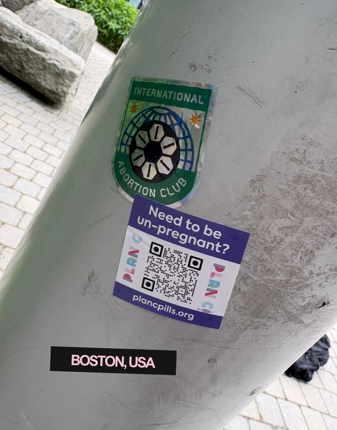 A sticker with the logo of the International Abortion Club on a light-colored surface, along with a smaller sticker that asks, 'Need to be un-pregnant?' with QR codes and the website planpills.org. The location tag says Boston, USA.