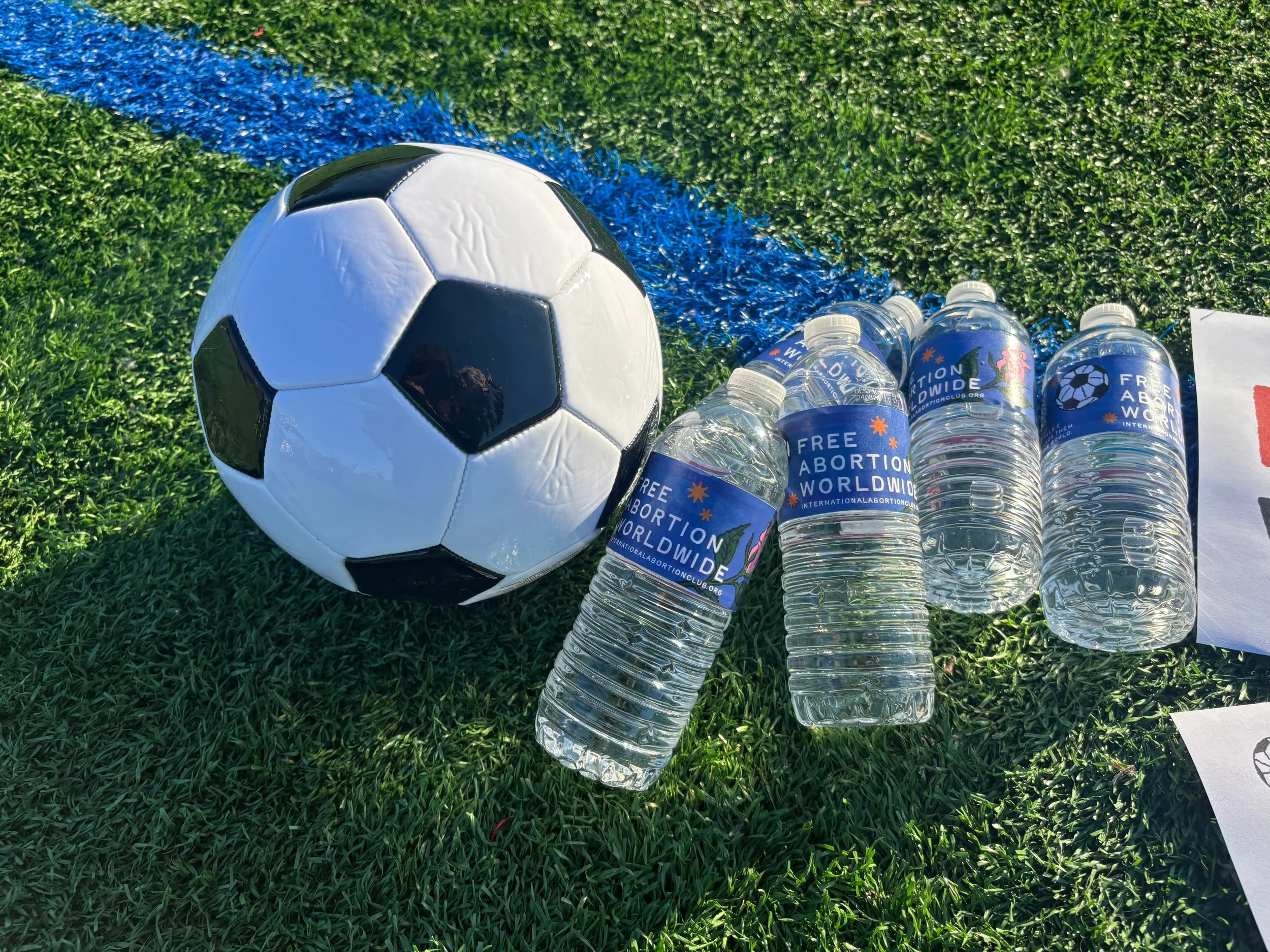 A soccer ball sits on green artificial turf next to several bottles of water with labels advocating for free abortion worldwide, along with a white piece of paper.
