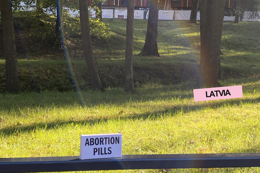 A grassy outdoor area with trees, a rainbow, and sunlight. There are two signs: one labeled "ABORTION PILLS" and another labeled "LATVIA."