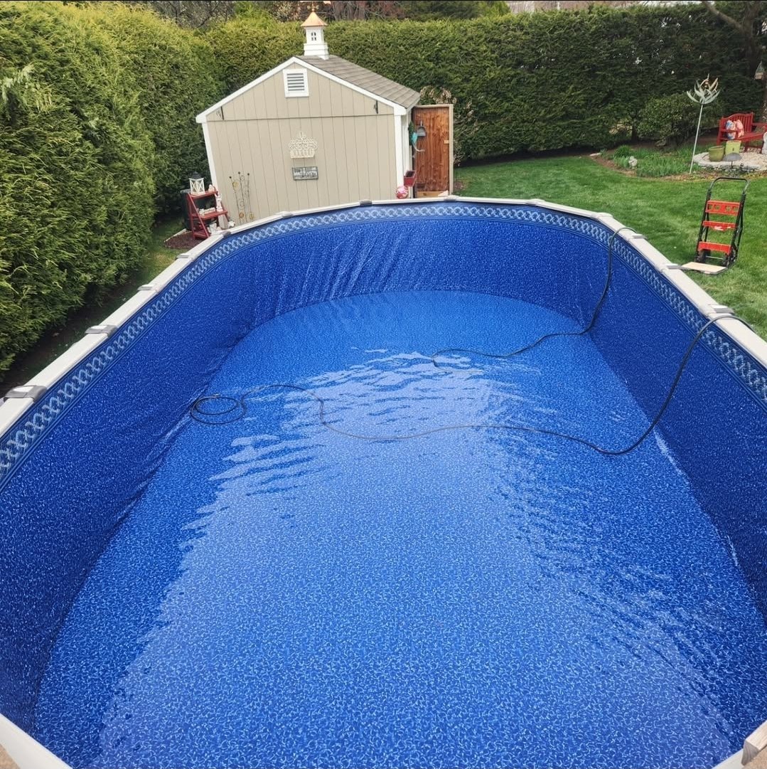 Above-ground backyard swimming pool with water. In the background, there is a shed, green hedges, and a grassy yard with outdoor furniture and decorative items.