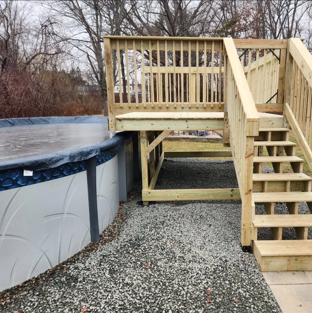 Wooden deck with stairs built next to an above-ground swimming pool in a backyard.
