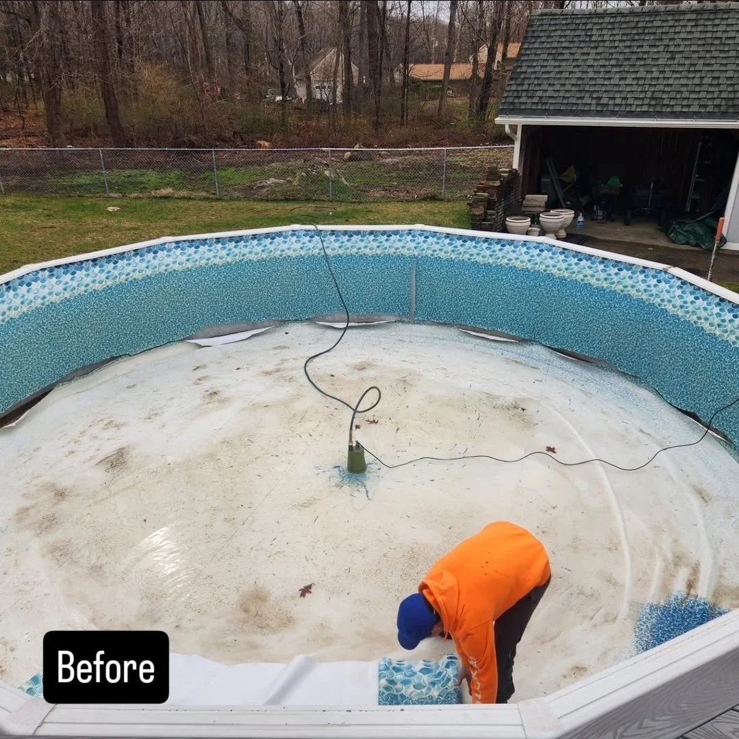 A person working inside an above-ground swimming pool that is empty and dirty, with a pool liner being installed. The yard has a fence, trees, a garage with gardening supplies, and a house in the background.