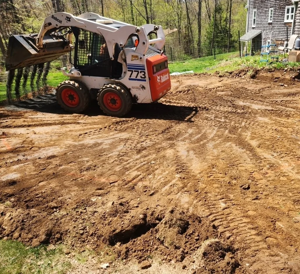 A Bobcat mini skid-steer loader with a digging bucket, parked on a cleared dirt area in a backyard, with a house and trees in the background.