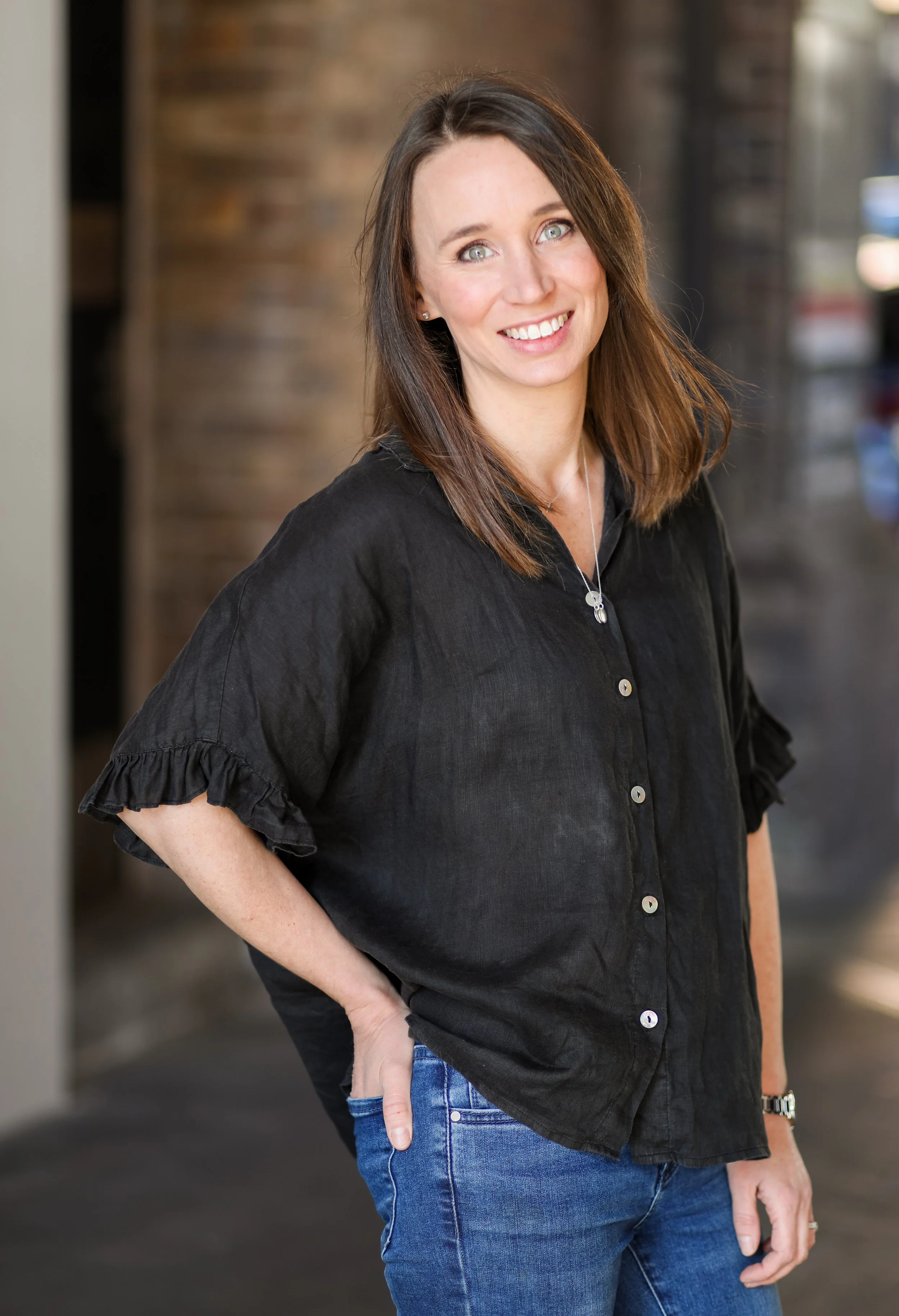 A woman with shoulder-length brown hair and blue eyes poses indoors against a blurred background of brown and gray tones, wearing a black shirt and blue jeans.