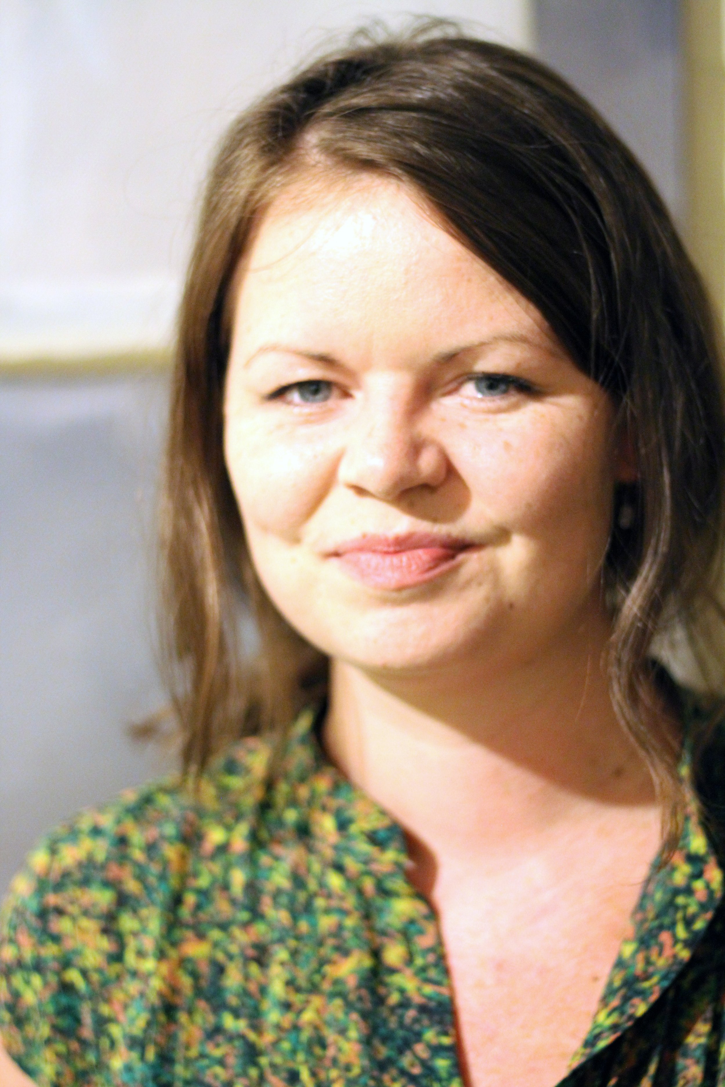A close-up of a woman with light skin, brown hair, and blue eyes, smiling slightly, wearing a multicolored floral print top.