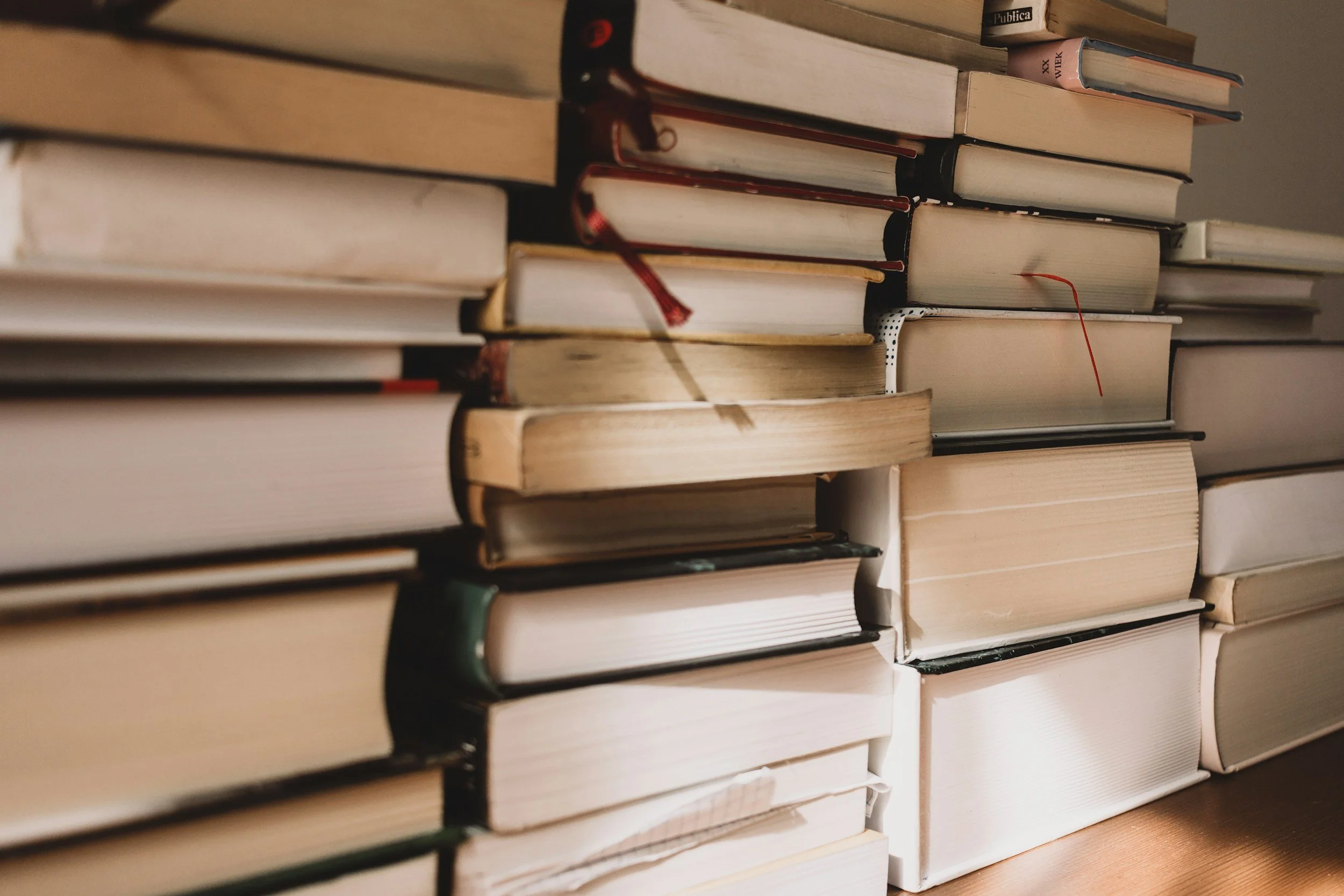 A close-up photo of a large stack of books with varying sizes and colors, resting on a wooden surface.