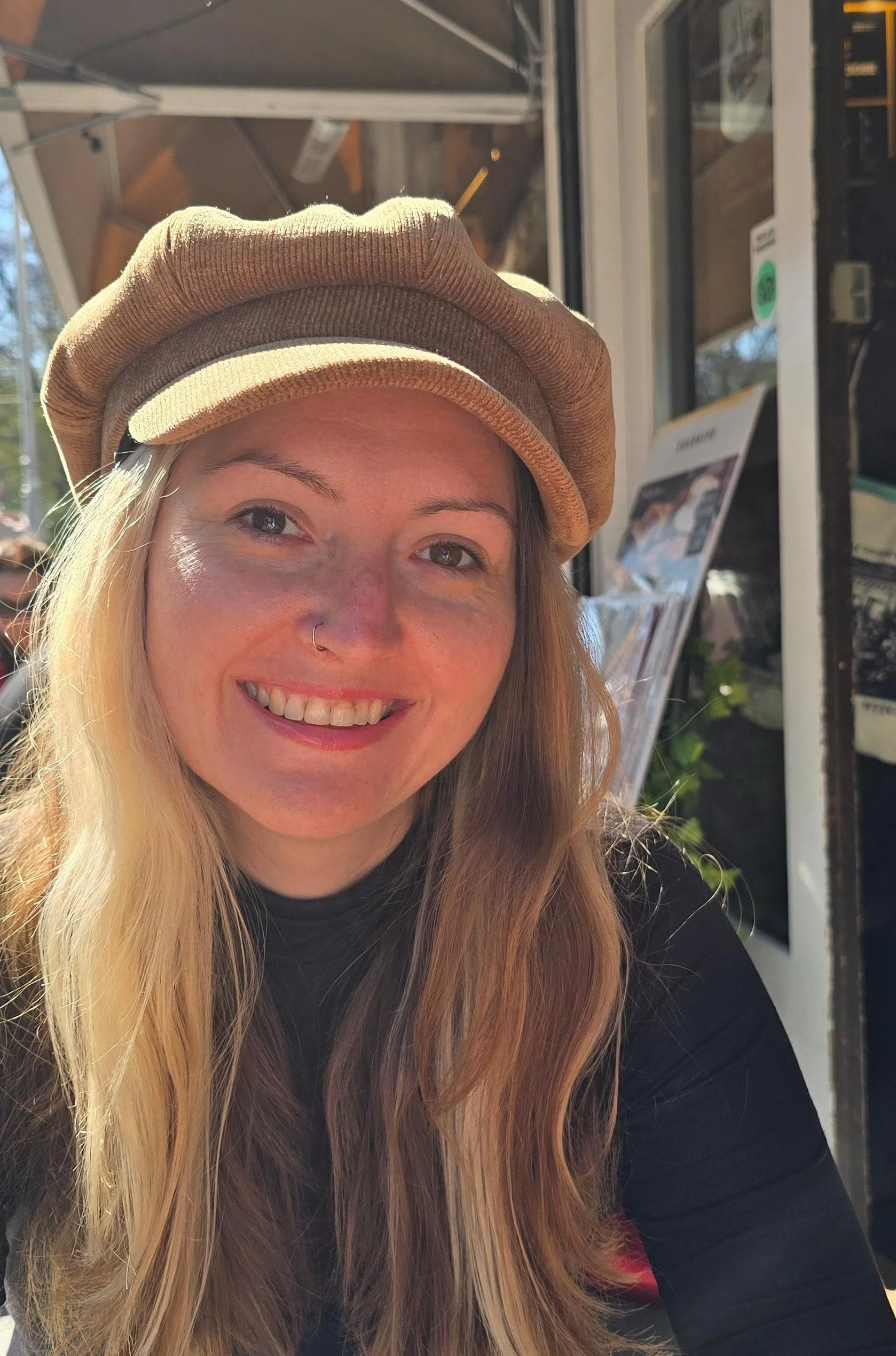 Close-up of a woman with long blonde and brown hair, smiling, wearing a tan cap and black top, sitting outside a cafe.