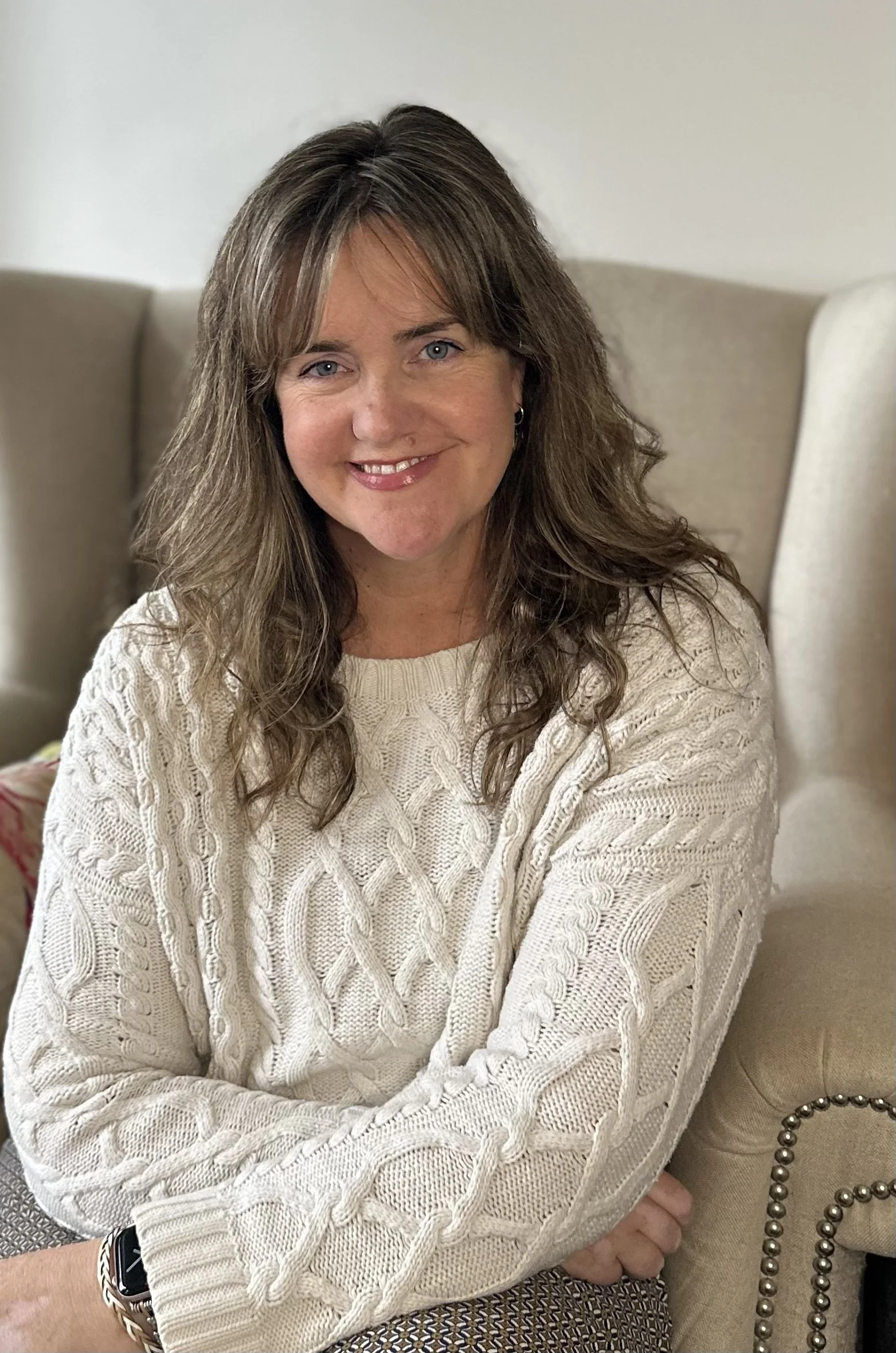 A woman with wavy brown hair and blue eyes, smiling, wearing a cream-colored cable knit sweater, sitting on a beige chair with a pearl embellishment, in a room with light-colored walls.