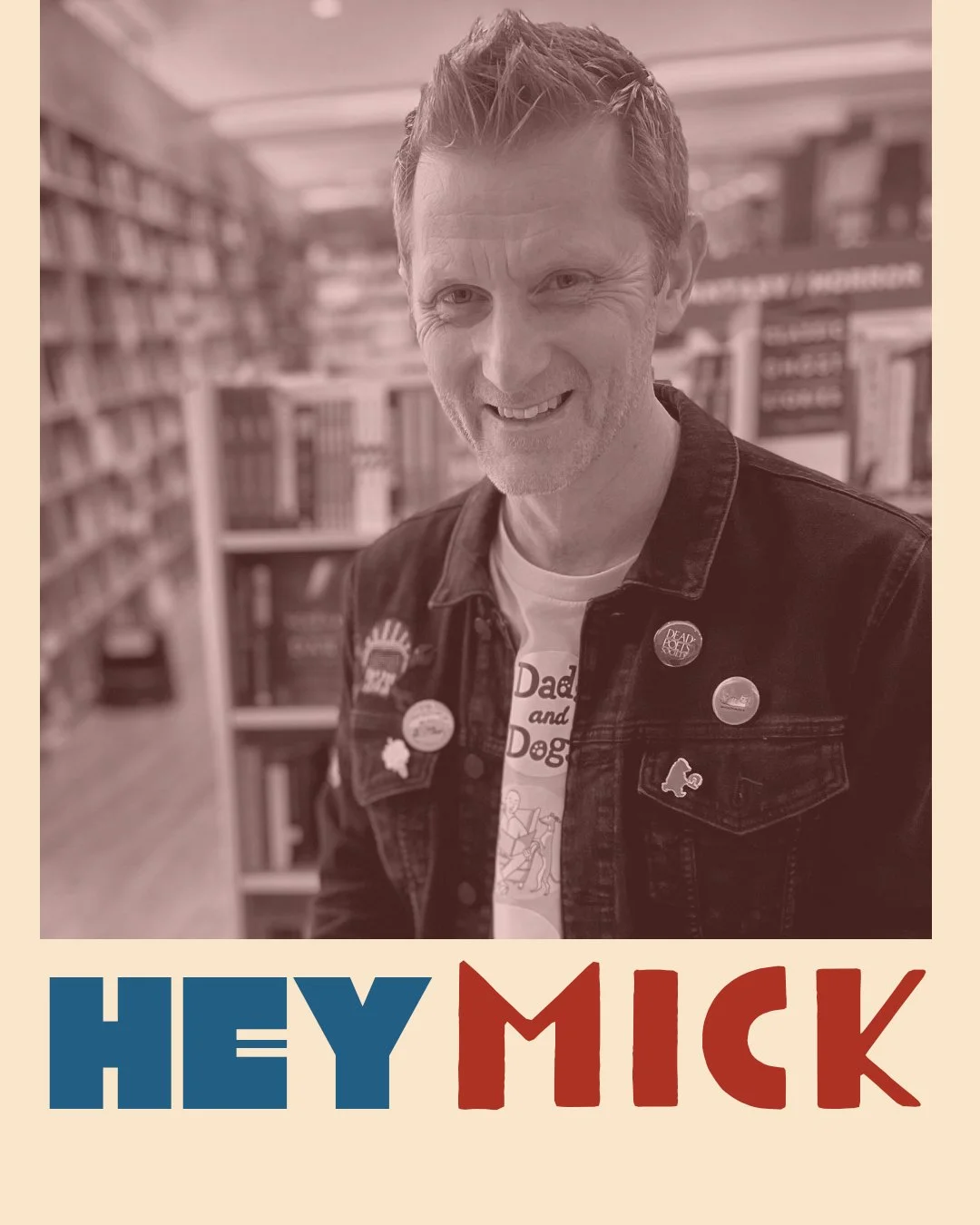 A smiling man with short hair and a beard, wearing a denim jacket with pins, in a bookstore with shelves of books in the background.