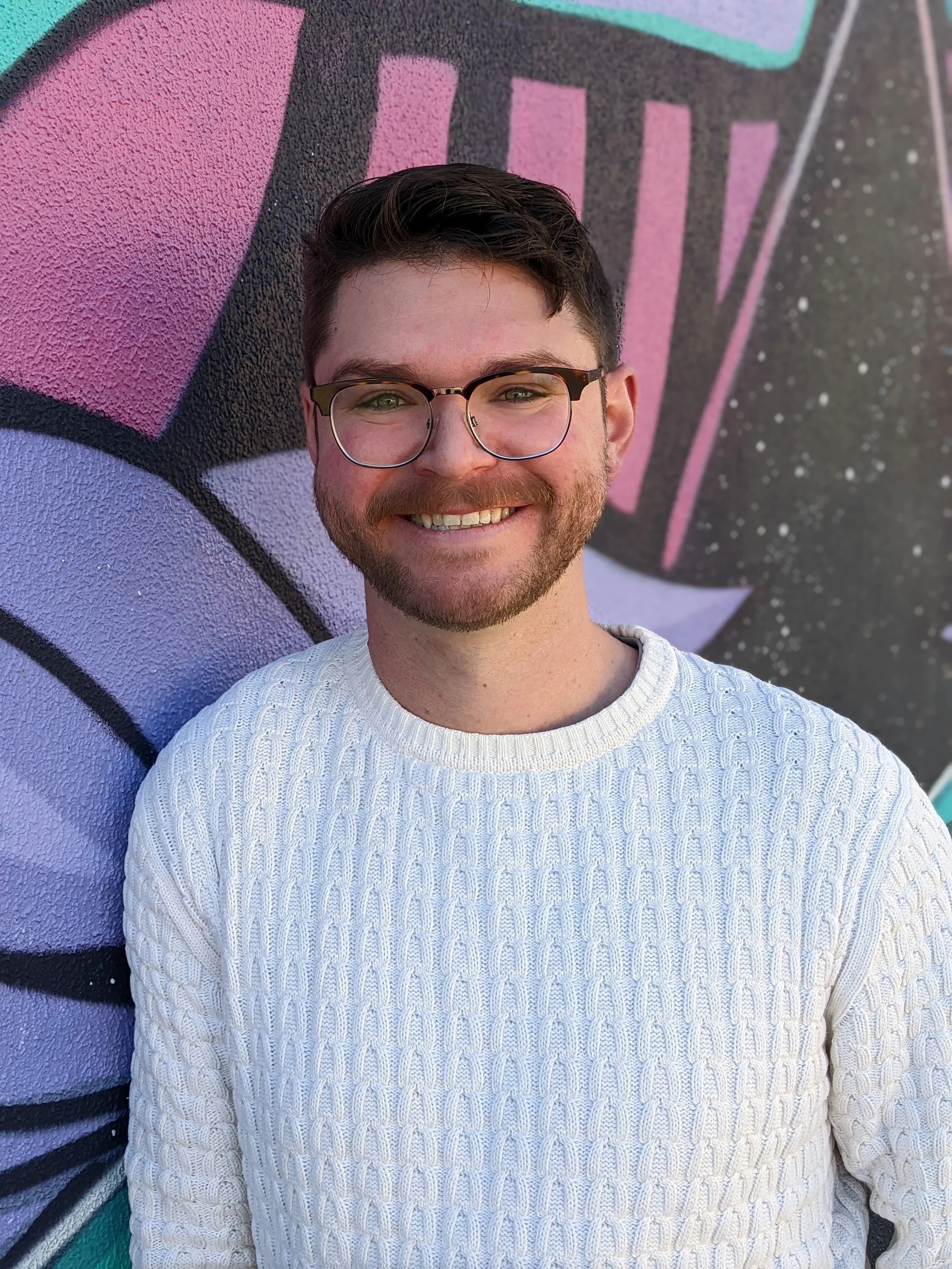 A man with glasses and a beard smiling, wearing a white sweater, standing in front of a pink and purple graffiti wall.