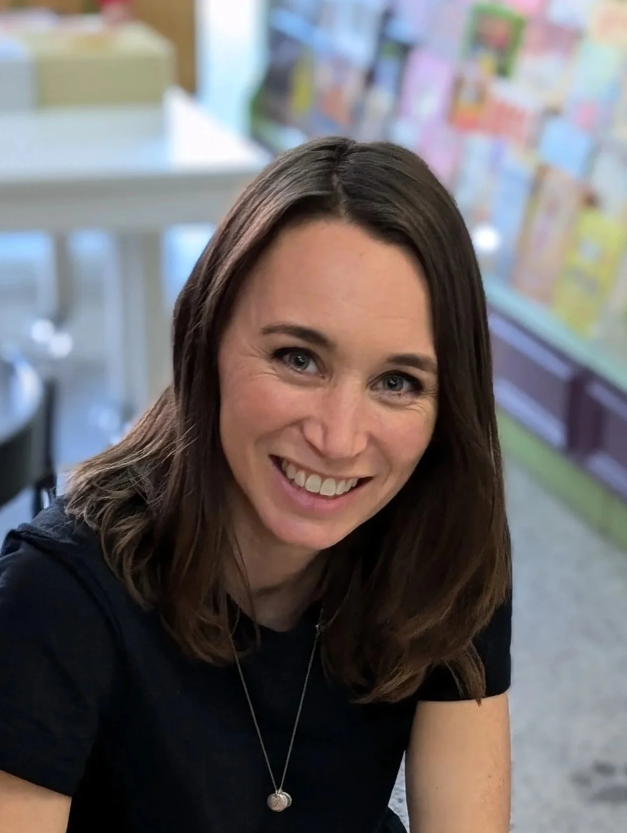 Close-up of a smiling woman with brown hair, wearing a black shirt and a silver necklace, in a room with blurred colorful posters or flyers in the background.
