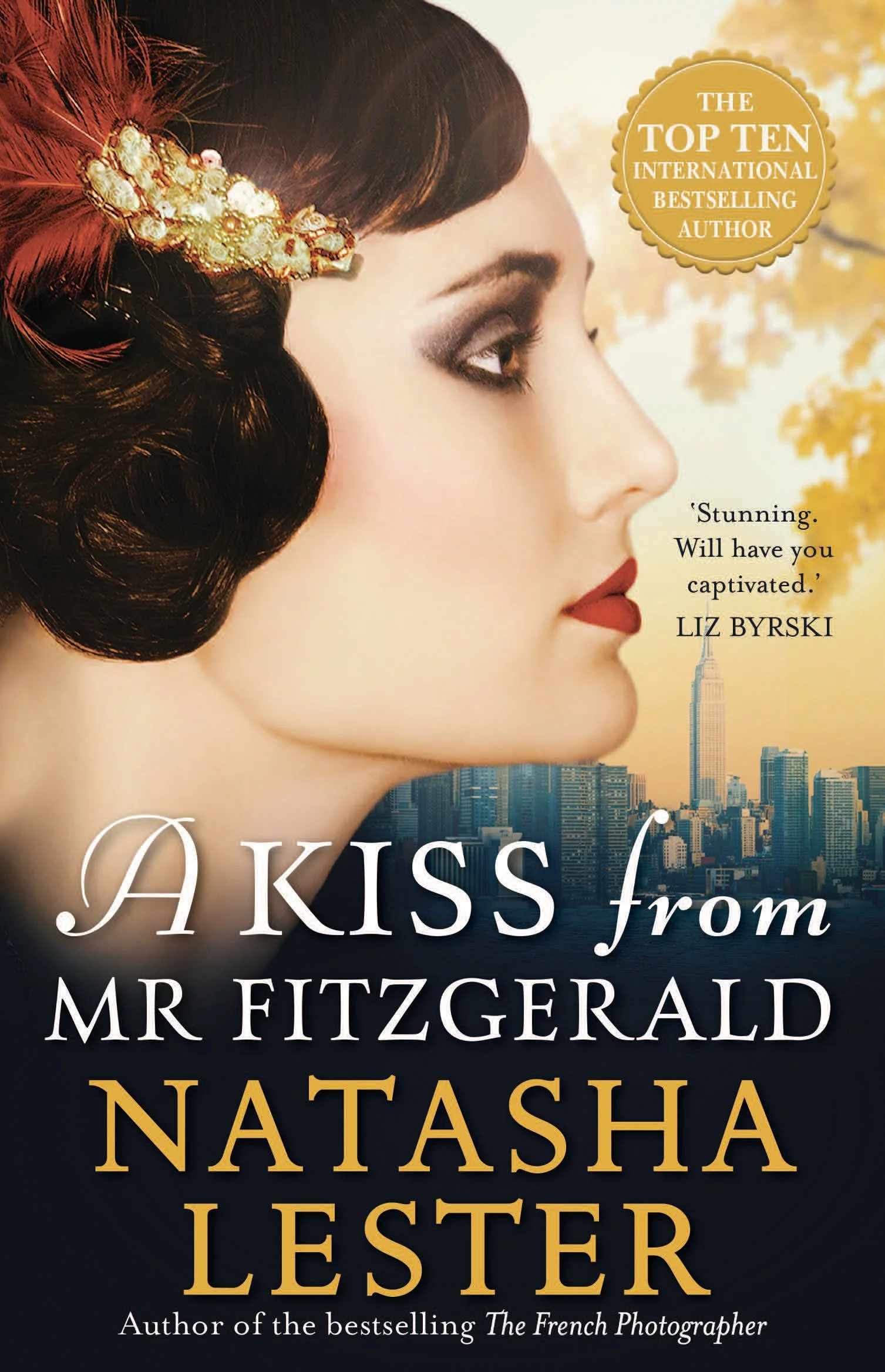 Book cover of 'A Kiss from Mr. Fitzgerald' by Natasha Lester featuring a side profile of a woman with vintage styling, dark hair, a decorative hairpiece with feathers and pearls, and a background of a city skyline, possibly New York City.