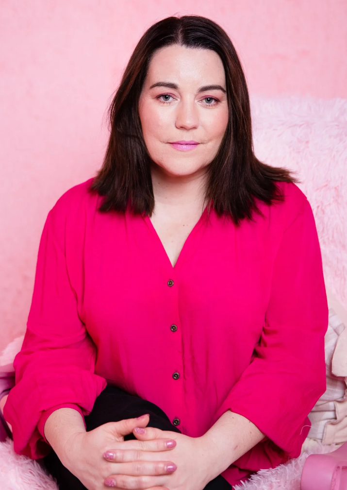 A woman with brown hair and fair skin wearing a bright pink blouse sitting on a soft pink fluffy surface against a pink background.