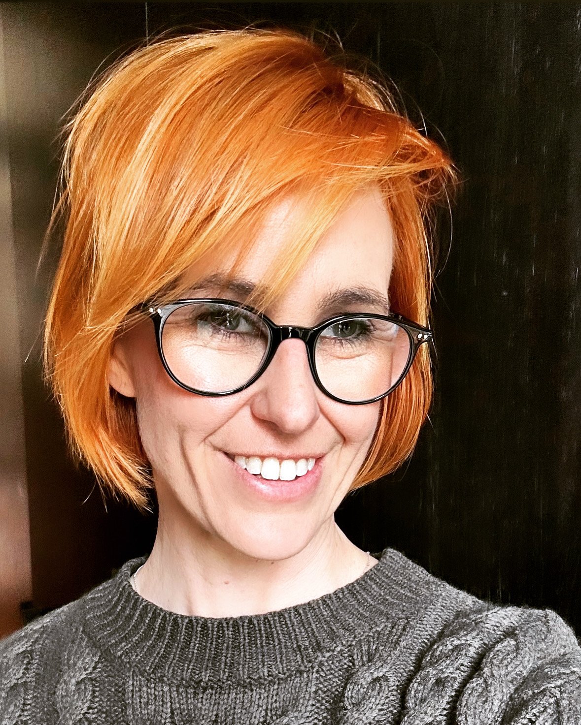 A woman with short, orange hair, wearing glasses and a gray knit sweater, smiling at the camera.