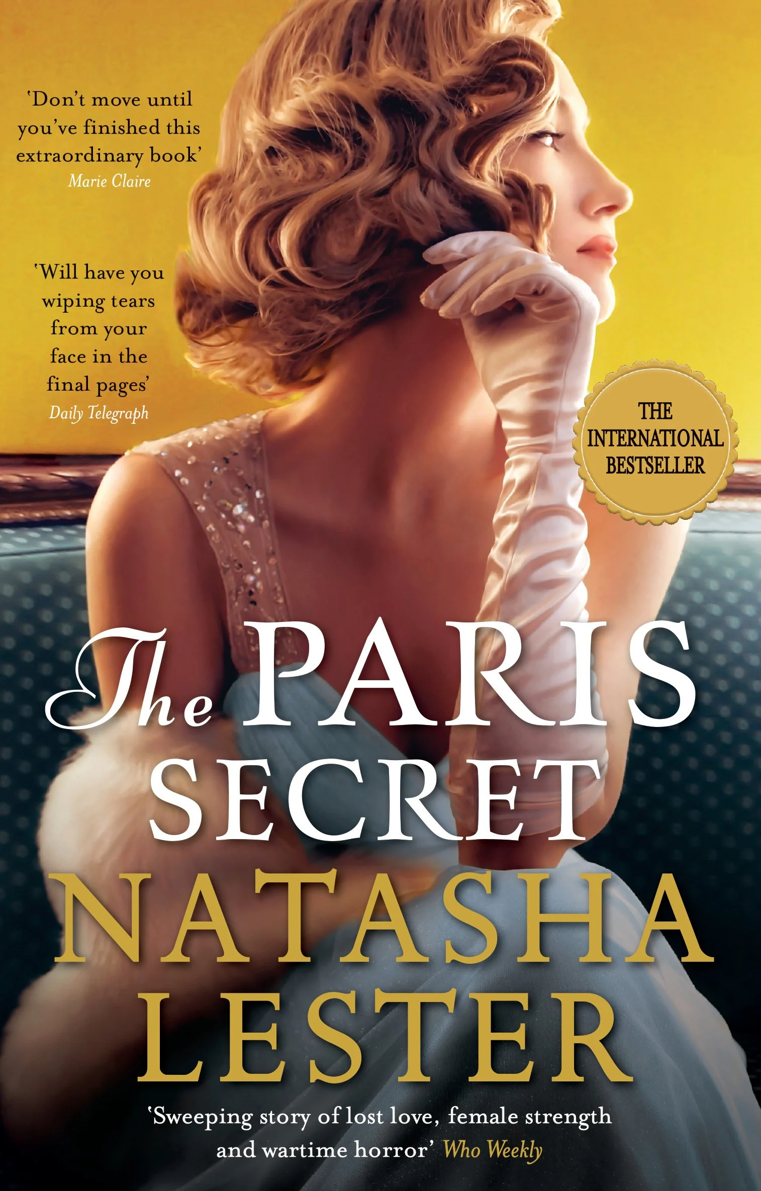 A woman with styled red hair, wearing a sleeveless dress with rhinestones, is seated indoors against a yellow background. She is touching her chin with her gloved hand, gazing to the right. The book cover features quotes and the title 'The Paris Secret' by Natasha Lester, with a gold sticker indicating it as a "The International Bestseller".