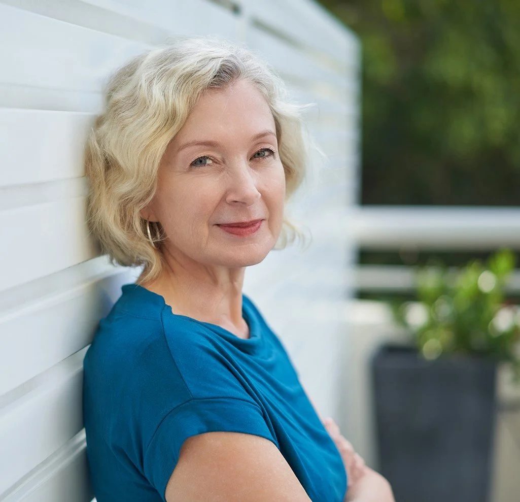 A middle-aged woman with blonde curly hair wearing a blue shirt, leaning against a white wall, smiling softly, with greenery in the background.