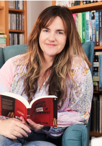 A woman with long brown hair is sitting on a blue armchair in a library, holding an open red book.