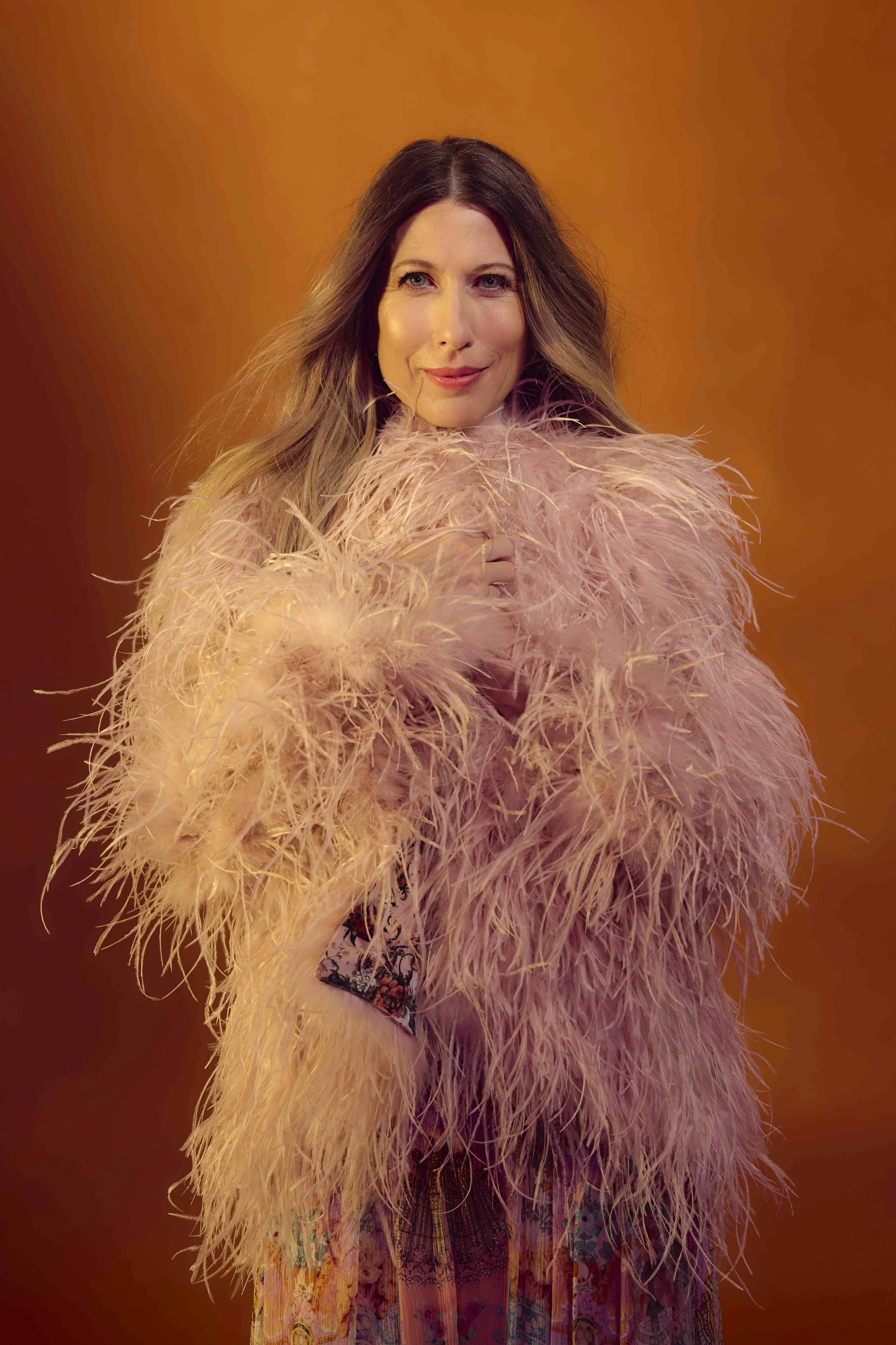 A woman with long brown hair wearing a pink feathered jacket over a floral dress, standing against an orange background.