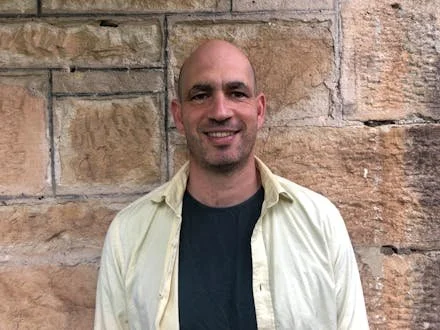 A man with a shaved head and light skin, smiling, wearing a light yellow shirt over a black T-shirt, standing in front of a stone wall.