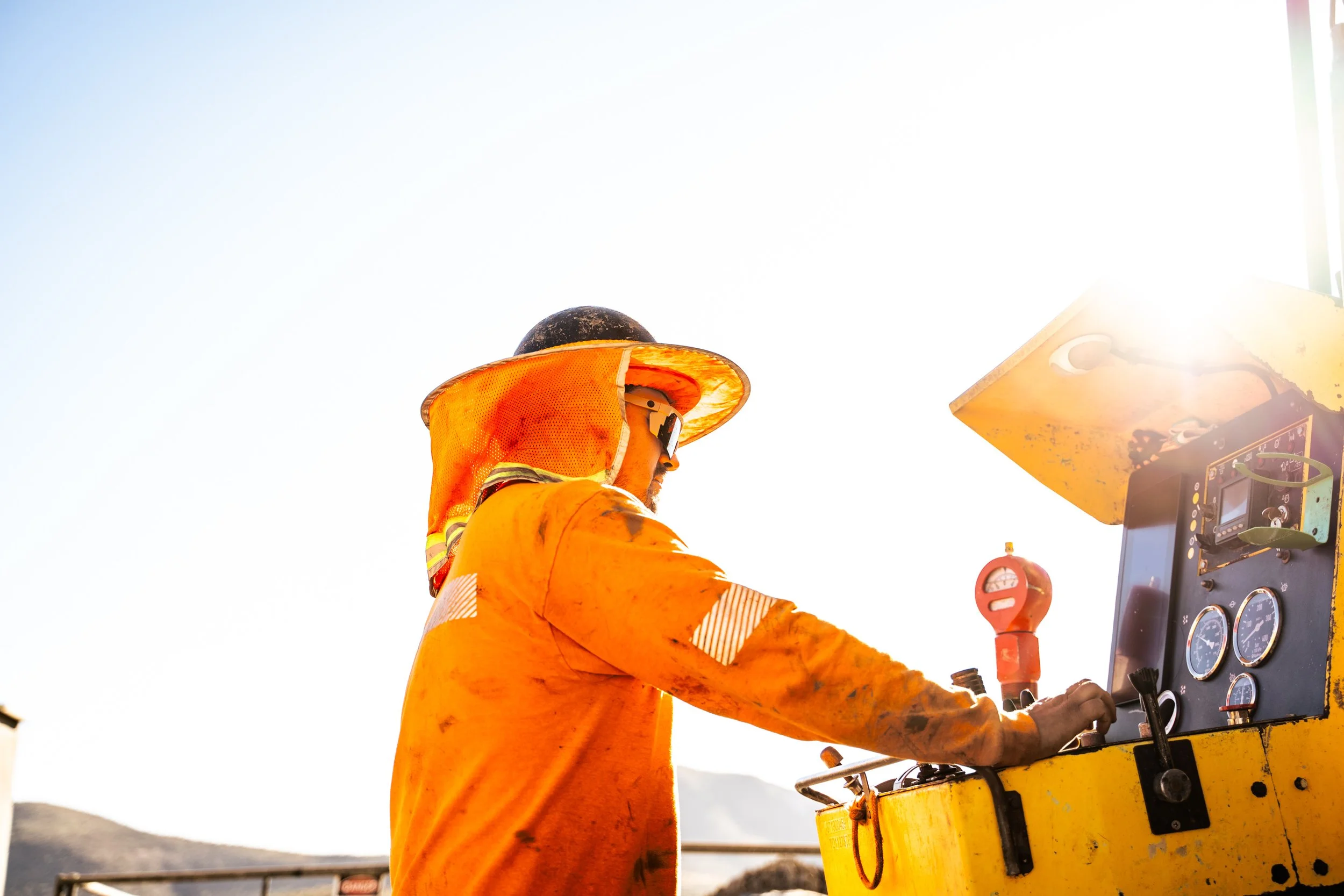 A construction worker in an orange safety jacket and helmet operating machinery outdoors in bright sunlight.
