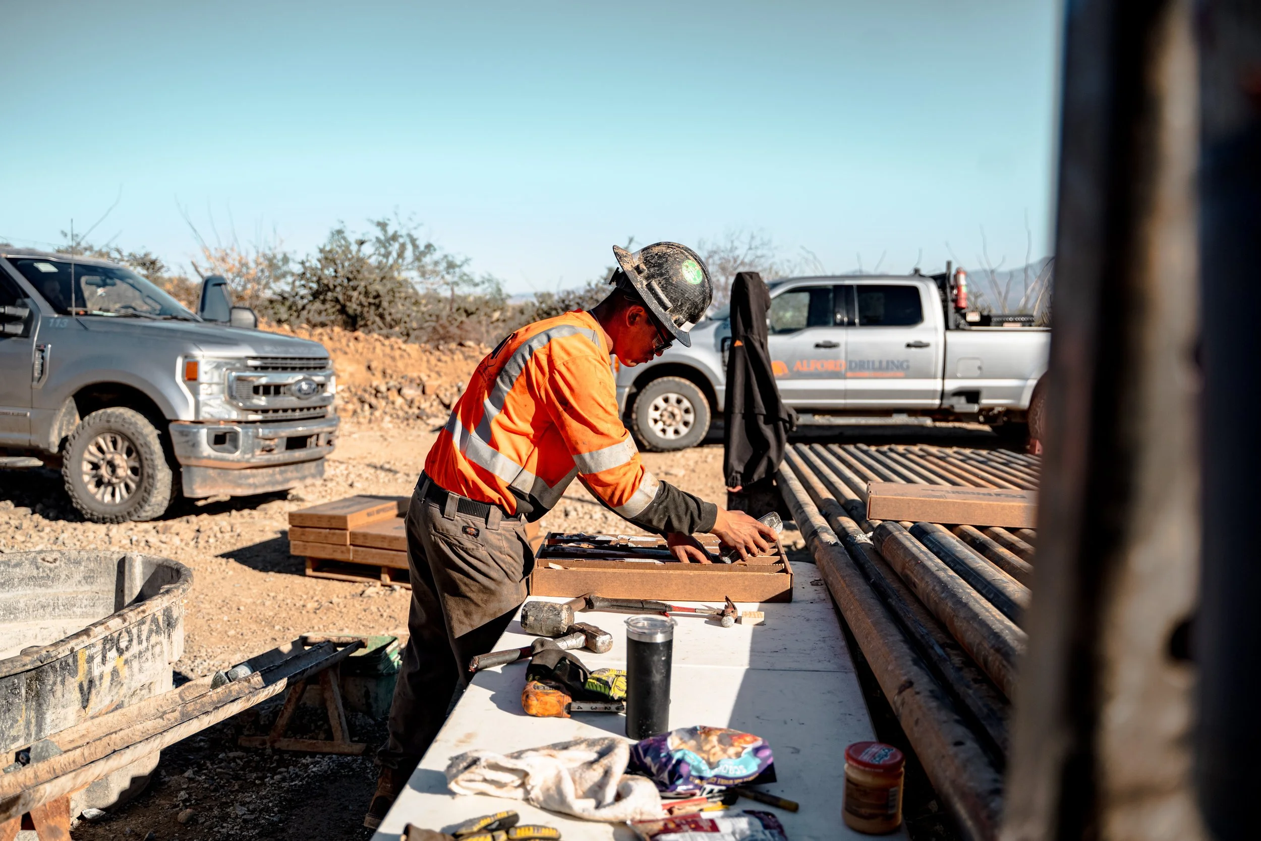 Construction worker in an orange safety vest and helmet arranging tools on a table at a construction site with trucks and equipment in the background.