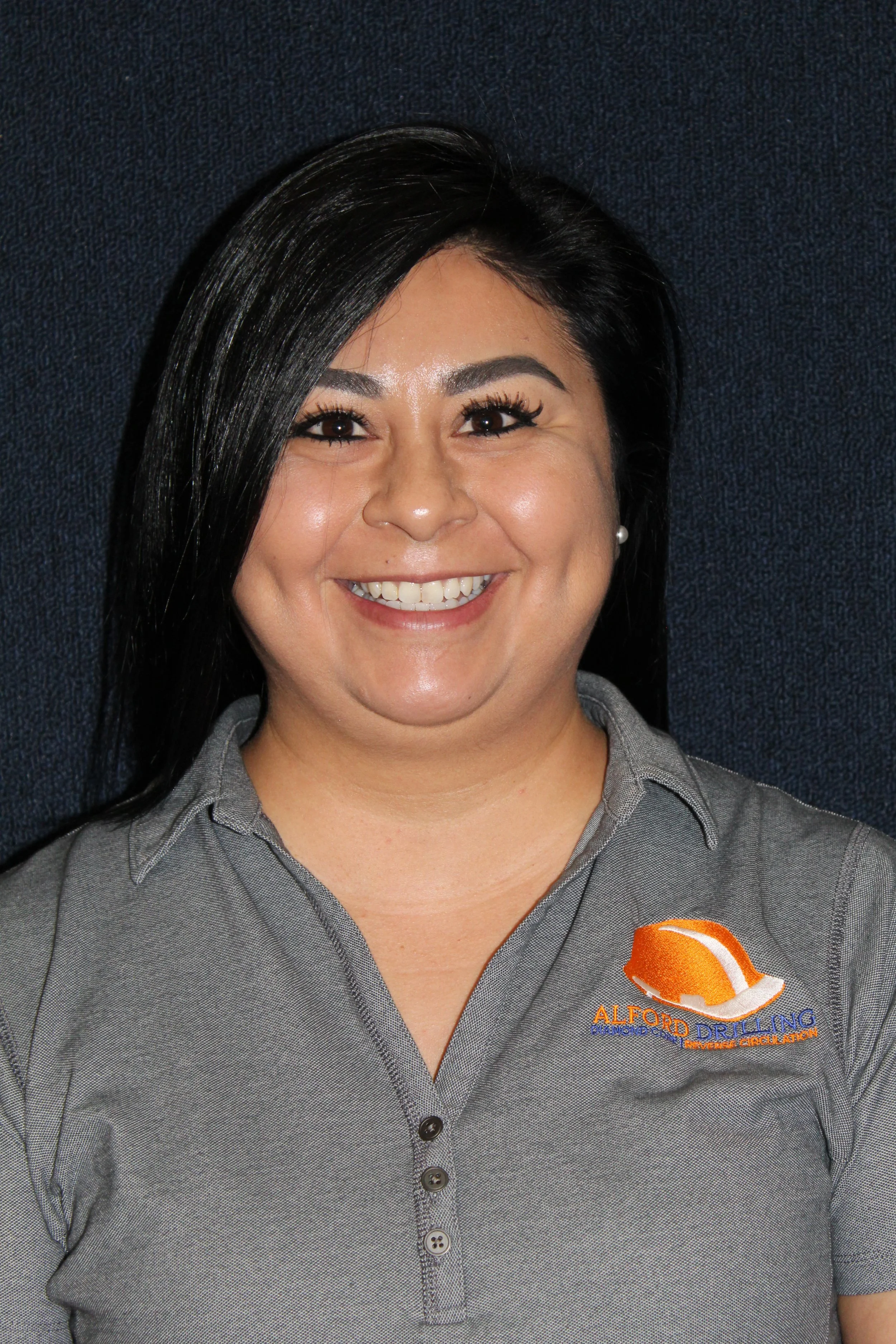 Smiling woman wearing a gray collared shirt with 'Alford Drilling' logo, dark hair, and pearl earrings, standing against a dark background.