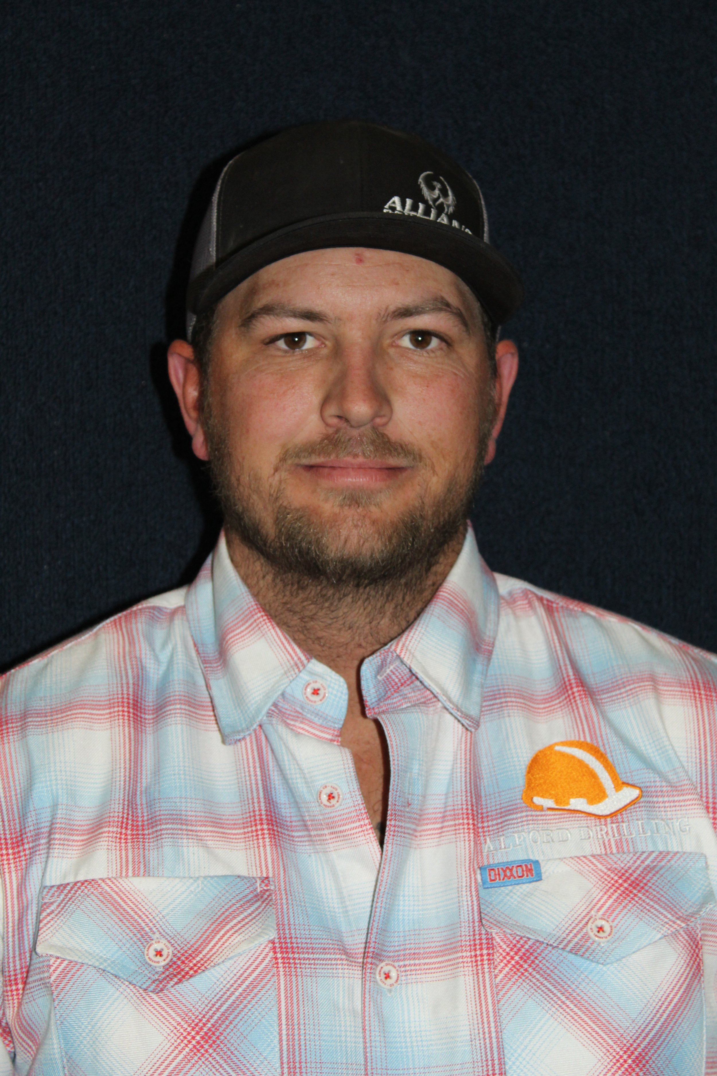 A man wearing a black baseball cap and a plaid button-up shirt with a logo, standing against a dark background.