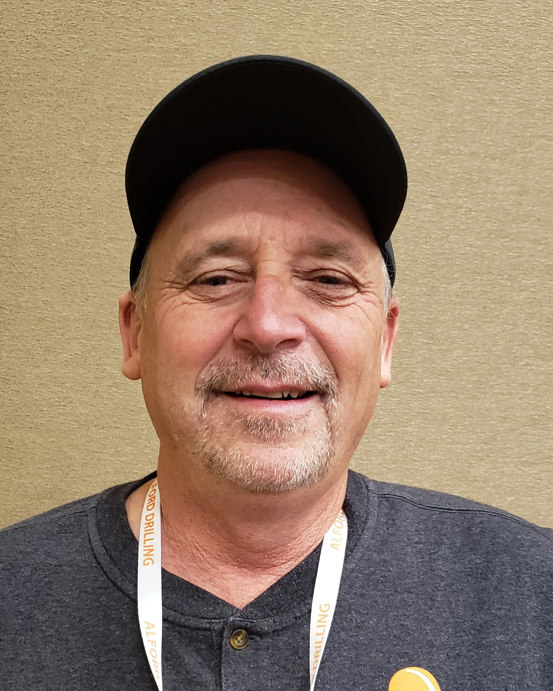 A close-up portrait of a smiling man with a beard, wearing a baseball cap and a lanyard, standing against a beige wall.