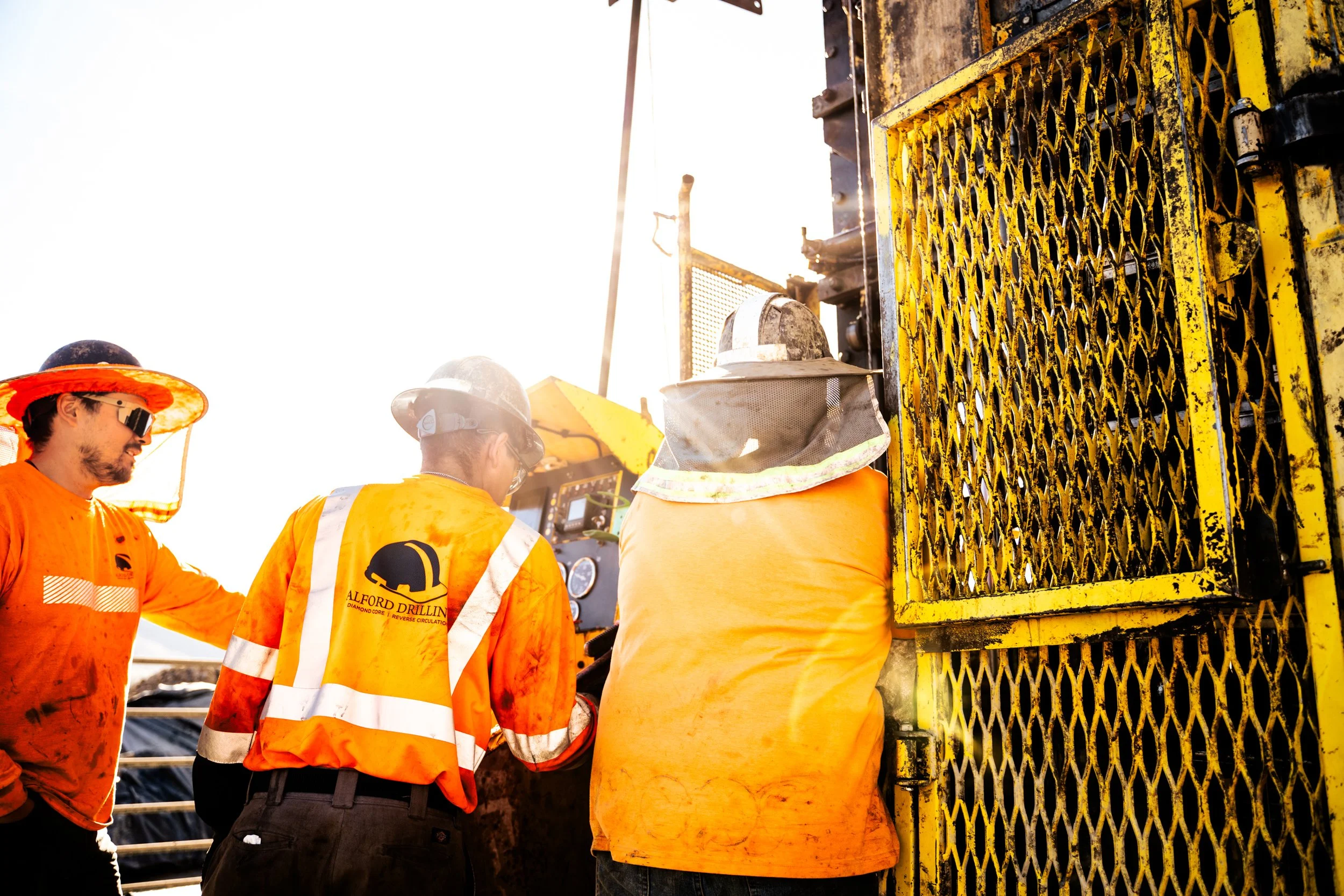 Three construction workers wearing safety gear, including helmets and high-visibility clothing, working on a drilling platform with machinery and yellow safety barriers, backlit by bright sunlight.