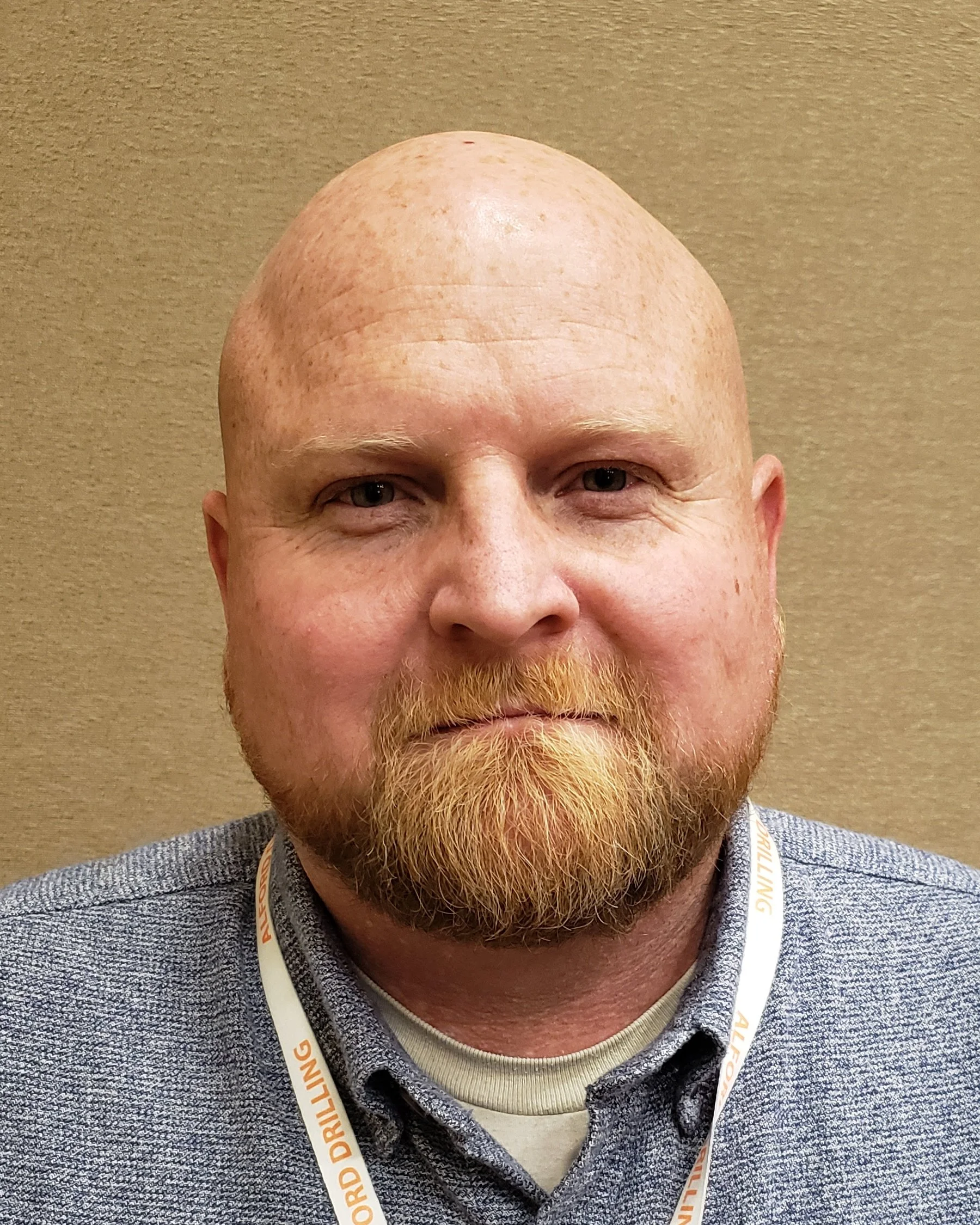 A close-up portrait of a bald man with a reddish beard, wearing a gray collared shirt and a white lanyard, standing against a beige background.