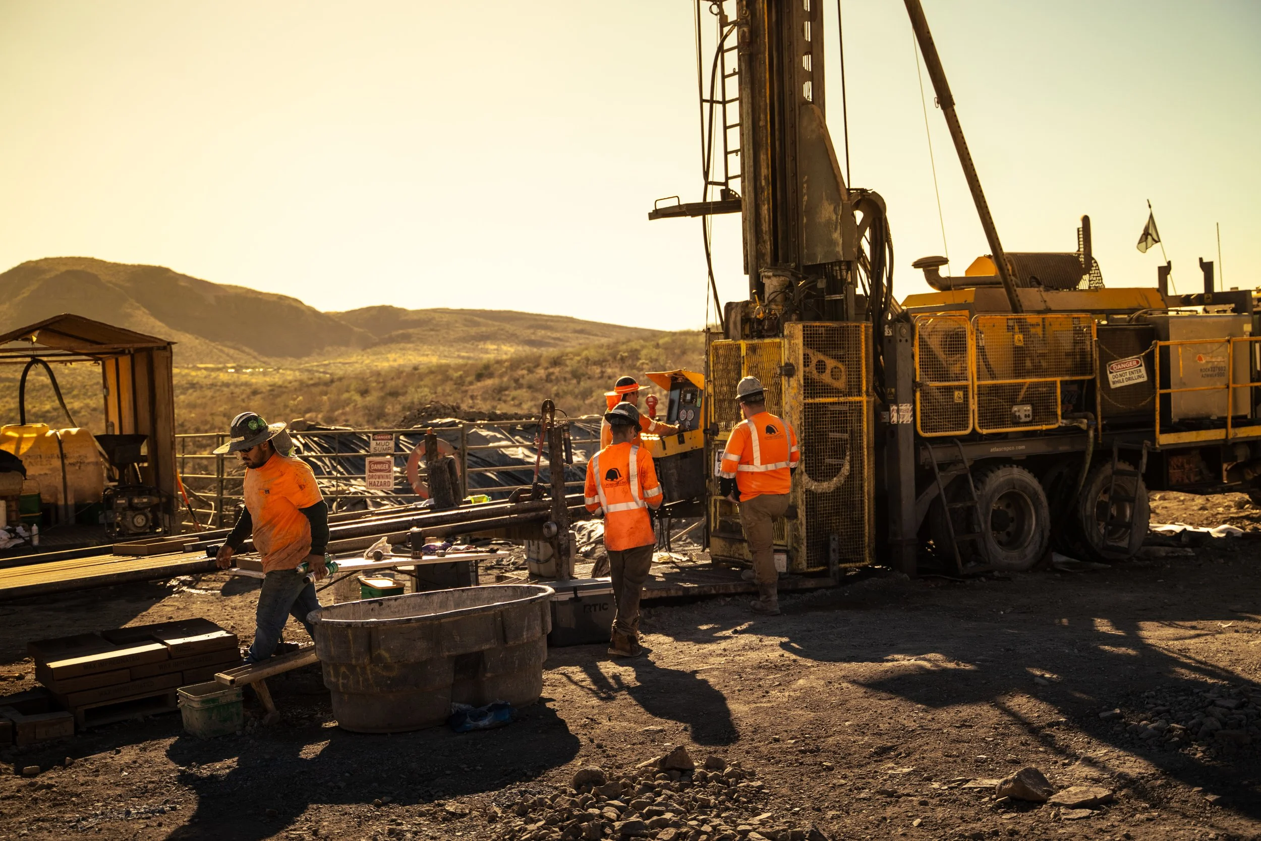 Construction workers operating heavy machinery on a construction site in a dry, mountainous landscape during sunset.