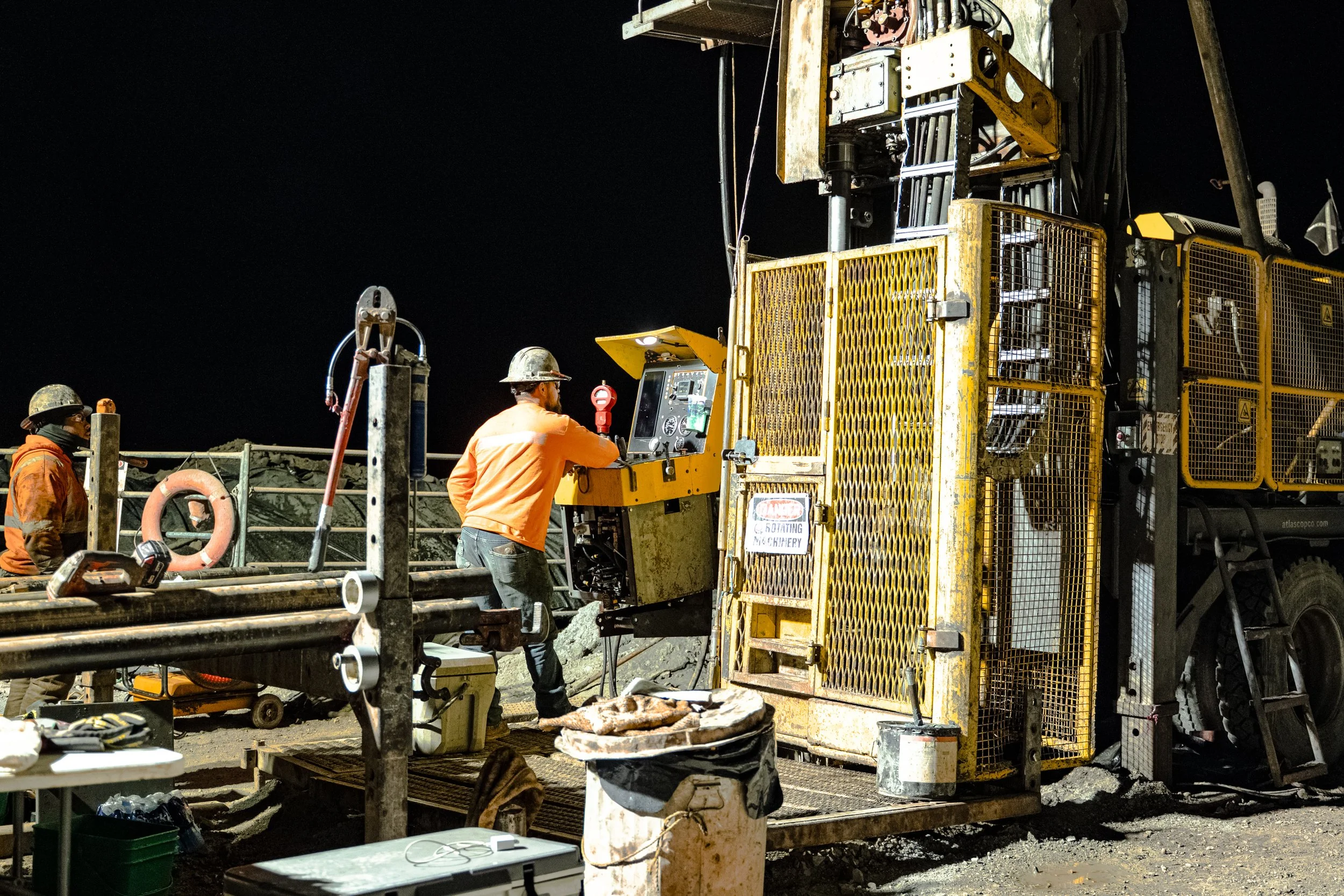 Workers operating heavy machinery at a construction site nighttime, with two workers in safety gear and helmets, one controlling a yellow drilling or blasting machine.