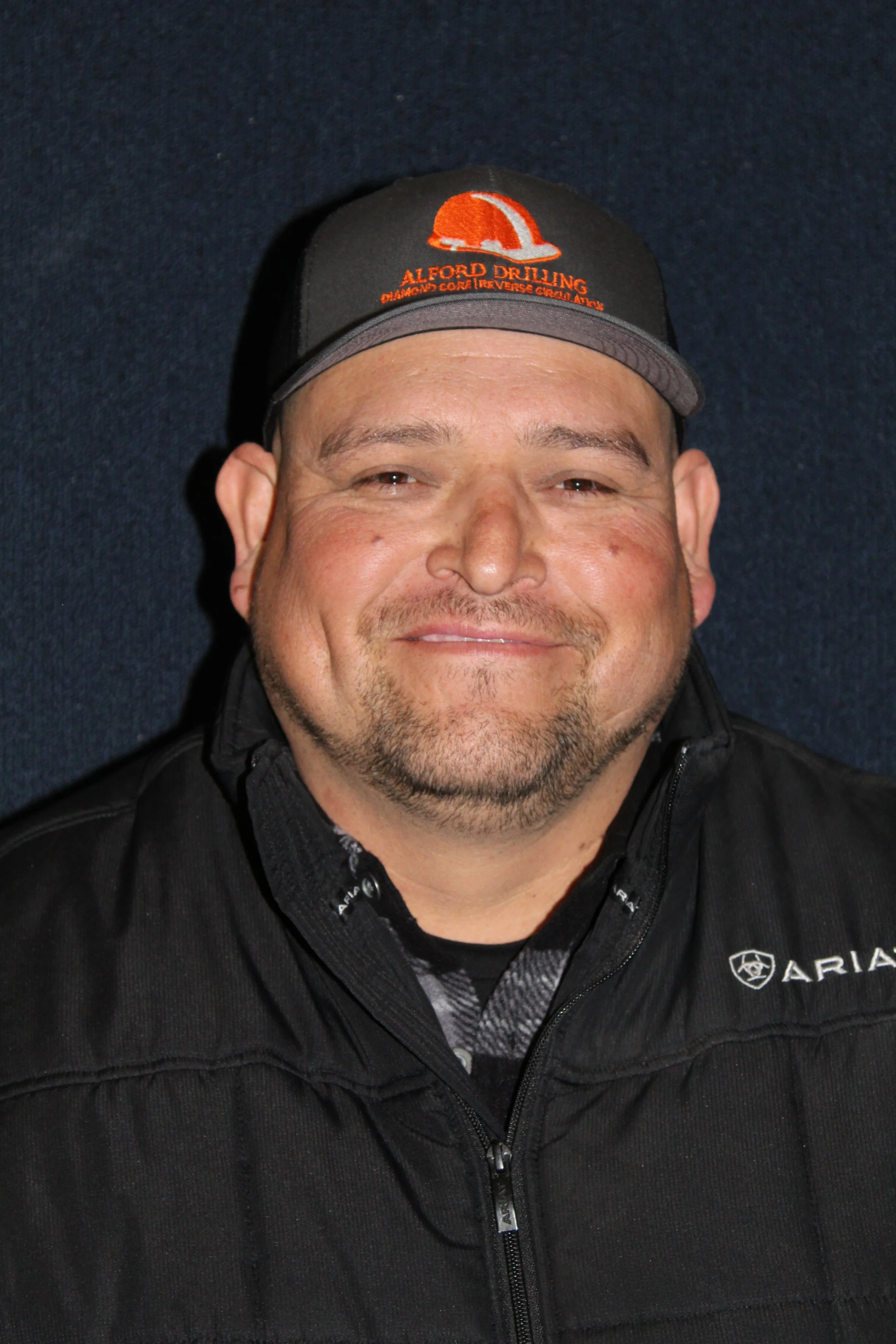 A man smiling wearing a black jacket and a black cap with orange and gray accents, with the logo and text 'Alford Drilling' on the cap, and the logo 'ARIA' on the jacket.