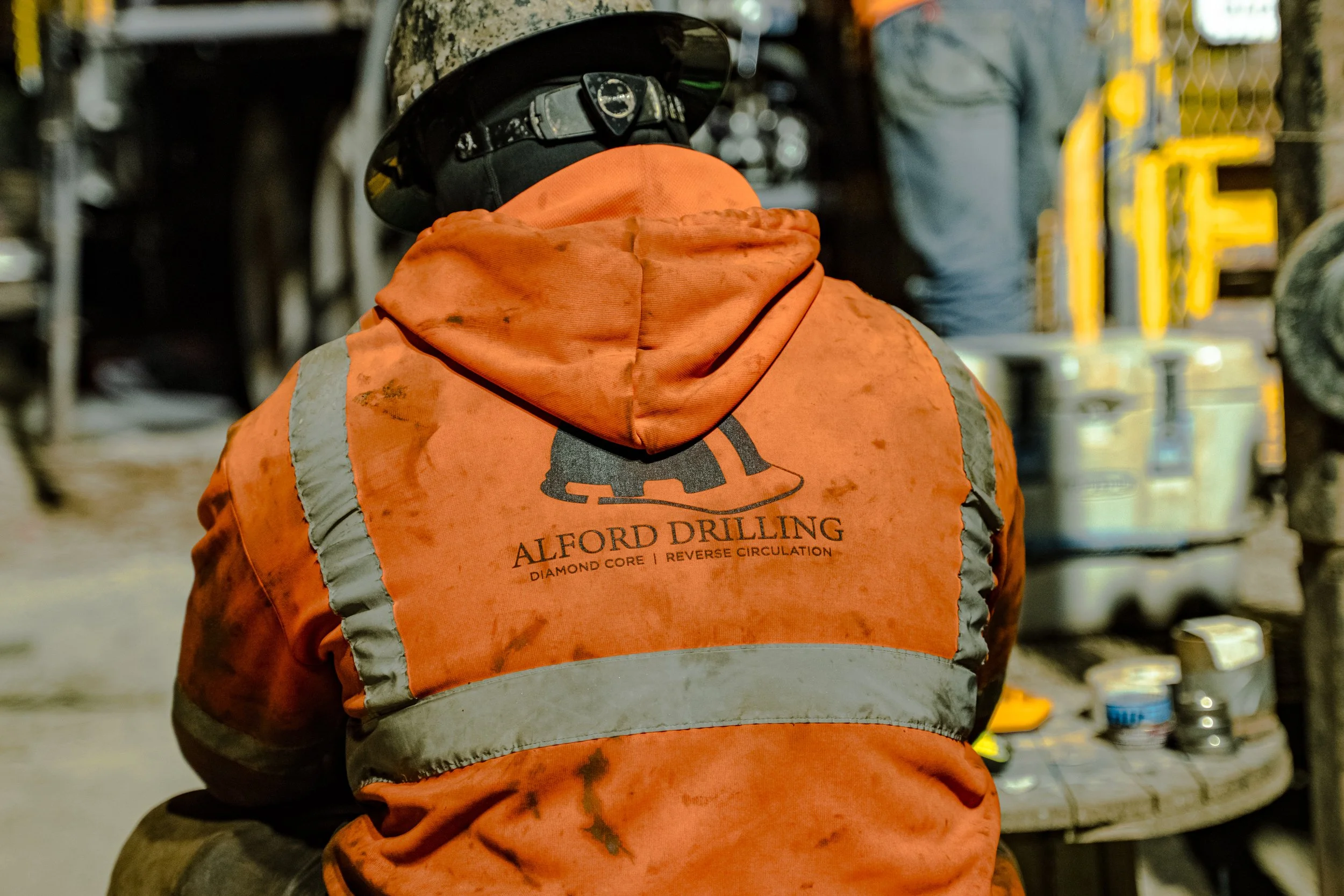 Construction worker wearing an orange jacket with 'Alford Drilling' logo, a black helmet, working at a cluttered workspace.