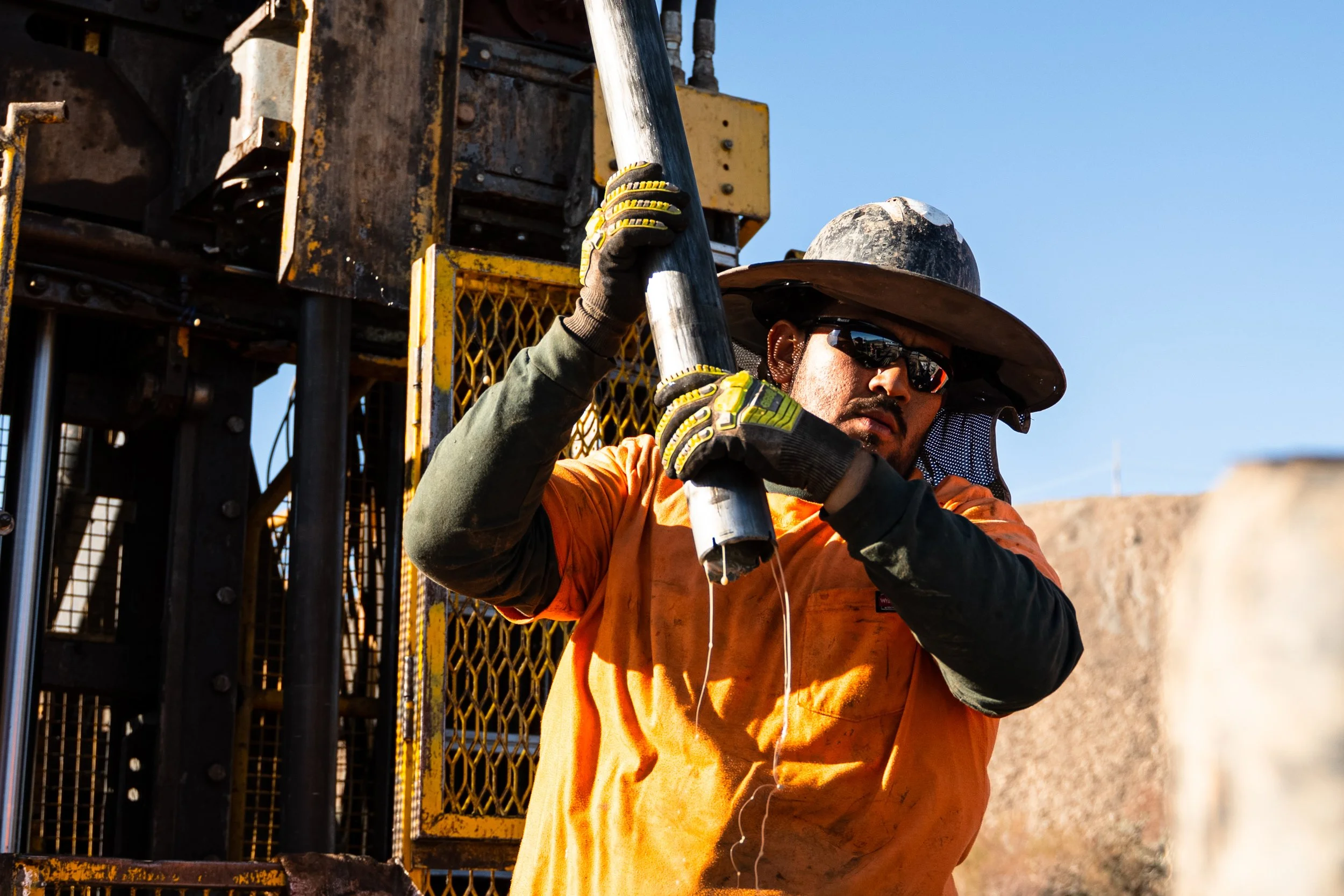 A construction worker wearing an orange safety shirt, black gloves, sunglasses, and a wide-brimmed hat is drilling into metal with a power tool at a construction site under clear blue skies.