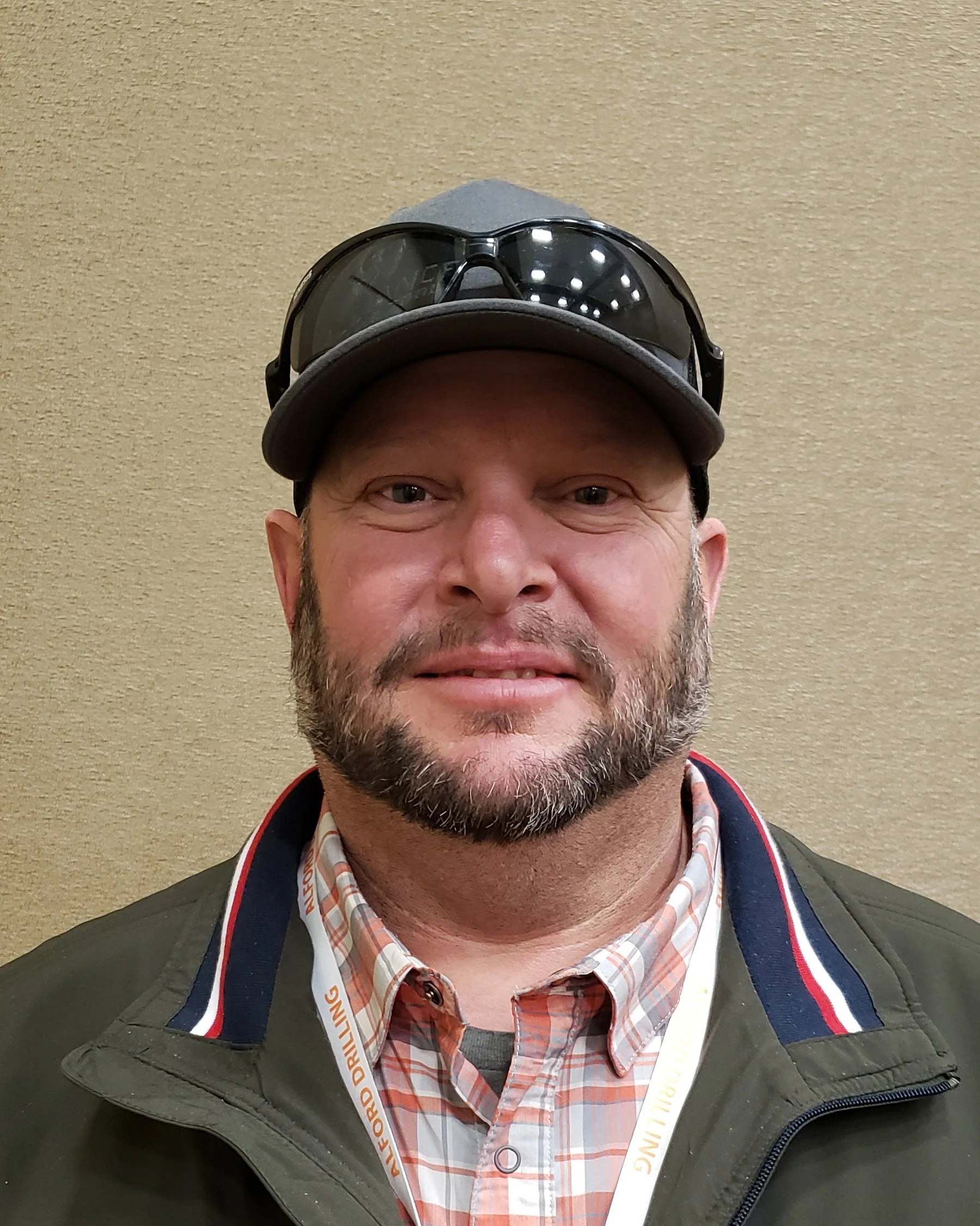 A man with a beard wearing a gray cap, dark sunglasses on top of the cap, a plaid shirt, and a dark jacket, standing against a beige wall.