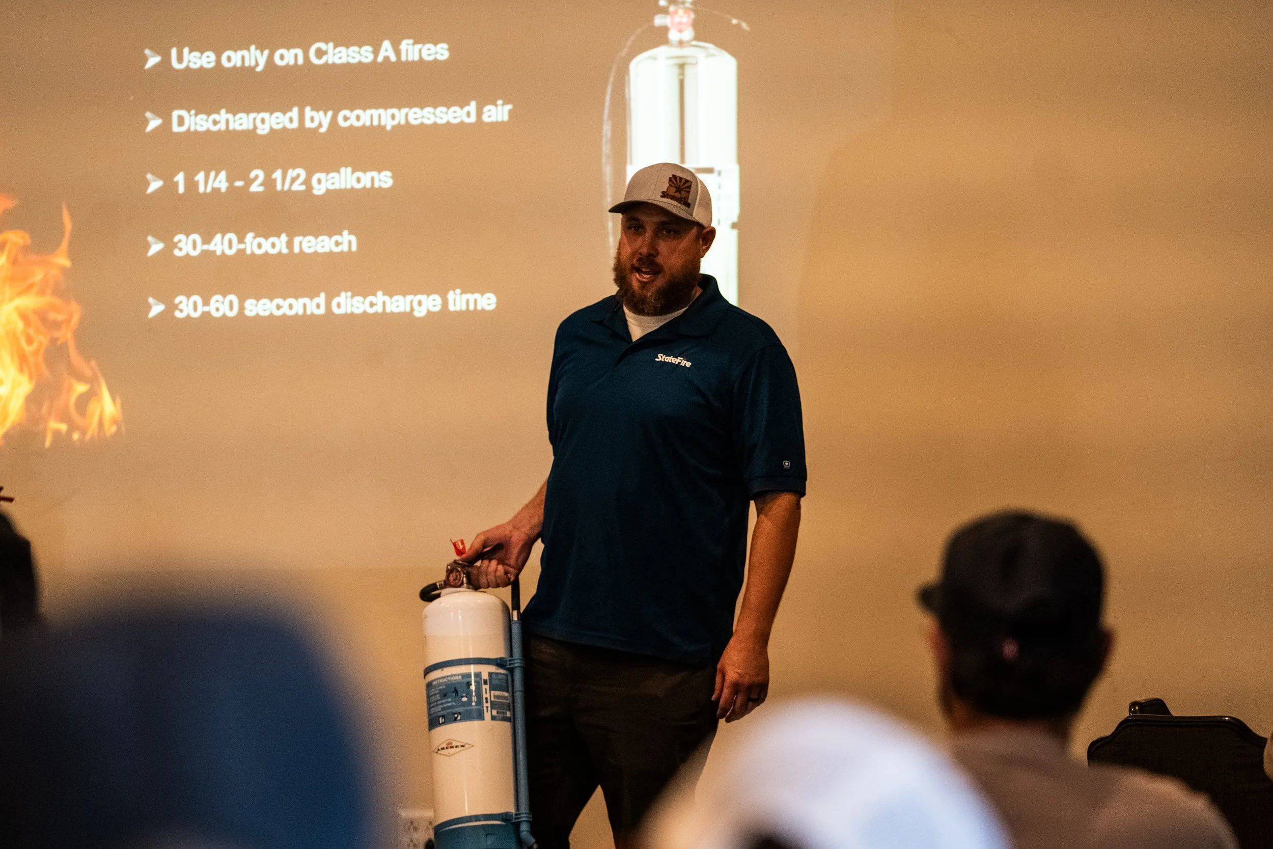 A man wearing a blue polo shirt and a baseball cap standing in front of a presentation slide about fire extinguishers, holding a fire extinguisher in his right hand. The slide lists instructions for using a fire extinguisher, including use only on Cl