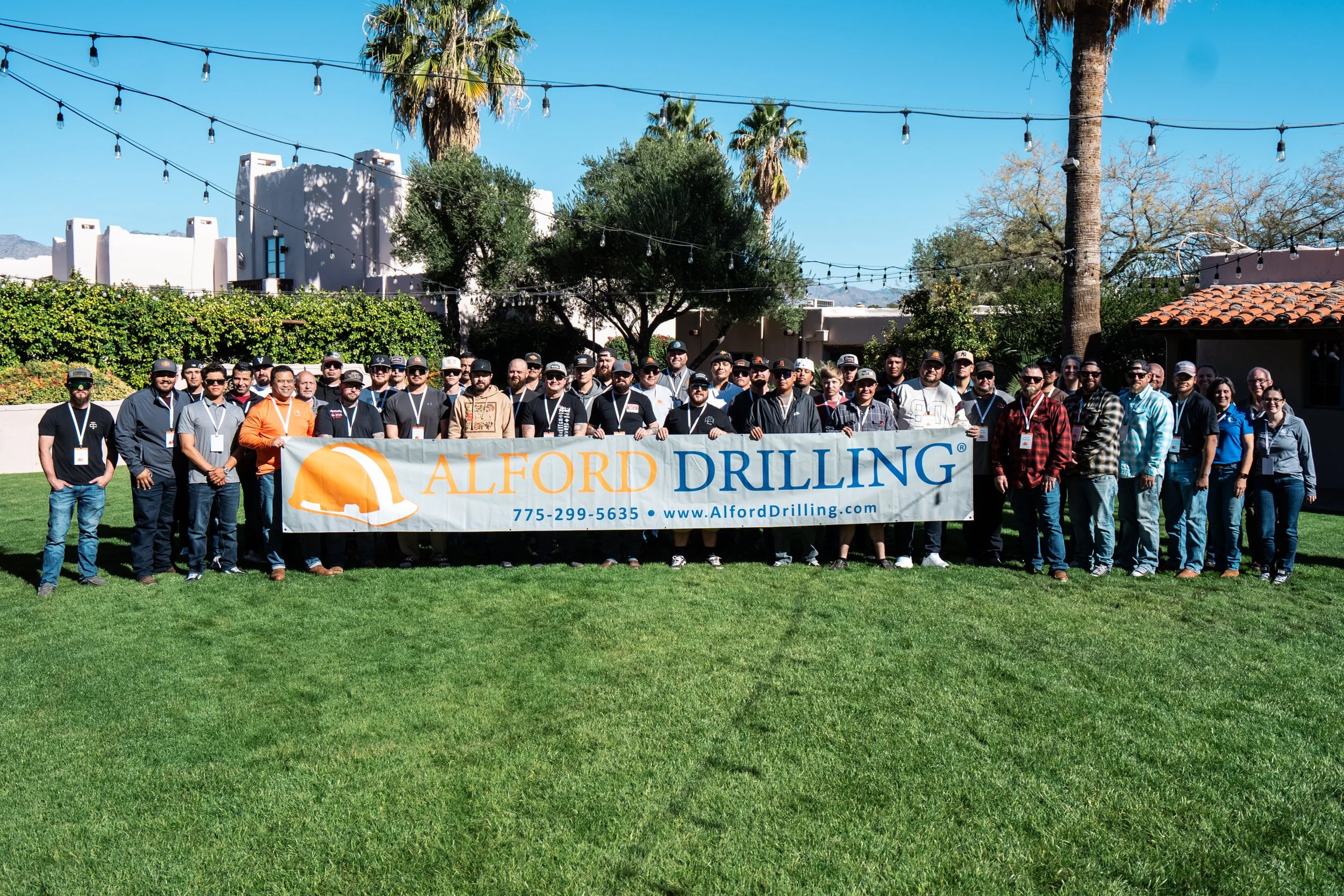 A large group of people standing outdoors on a grassy lawn holding a banner that reads "Alford Drilling" with a phone number and website. The background includes trees, bushes, a building, and string lights hanging above.