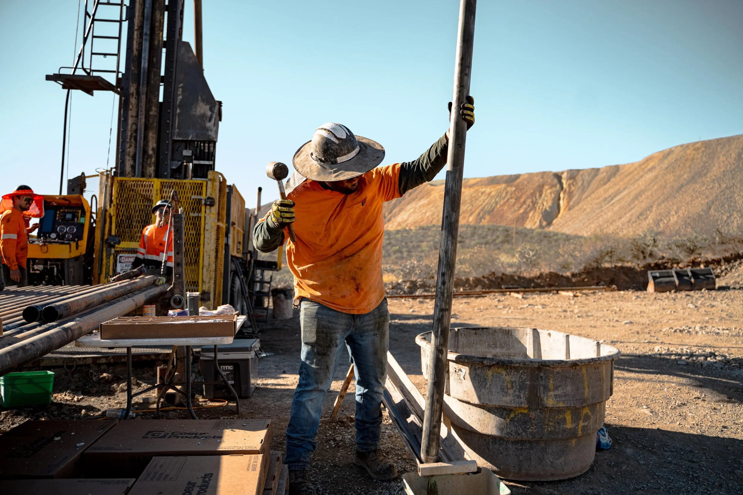 A construction worker in an orange shirt and wide-brimmed hat operates a large metal drill or pipe in an arid, rocky landscape with distant hills.