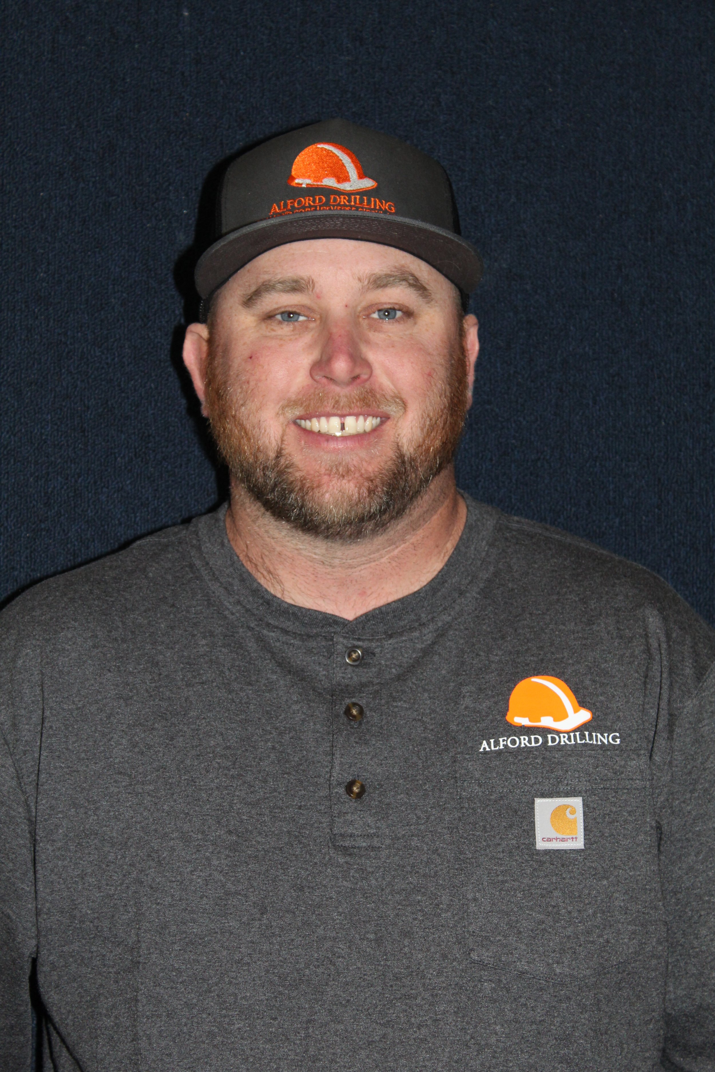 Man smiling wearing a gray Pedro shirt and a black cap with an orange construction helmet logo with the text 'Alford Drilling'.
