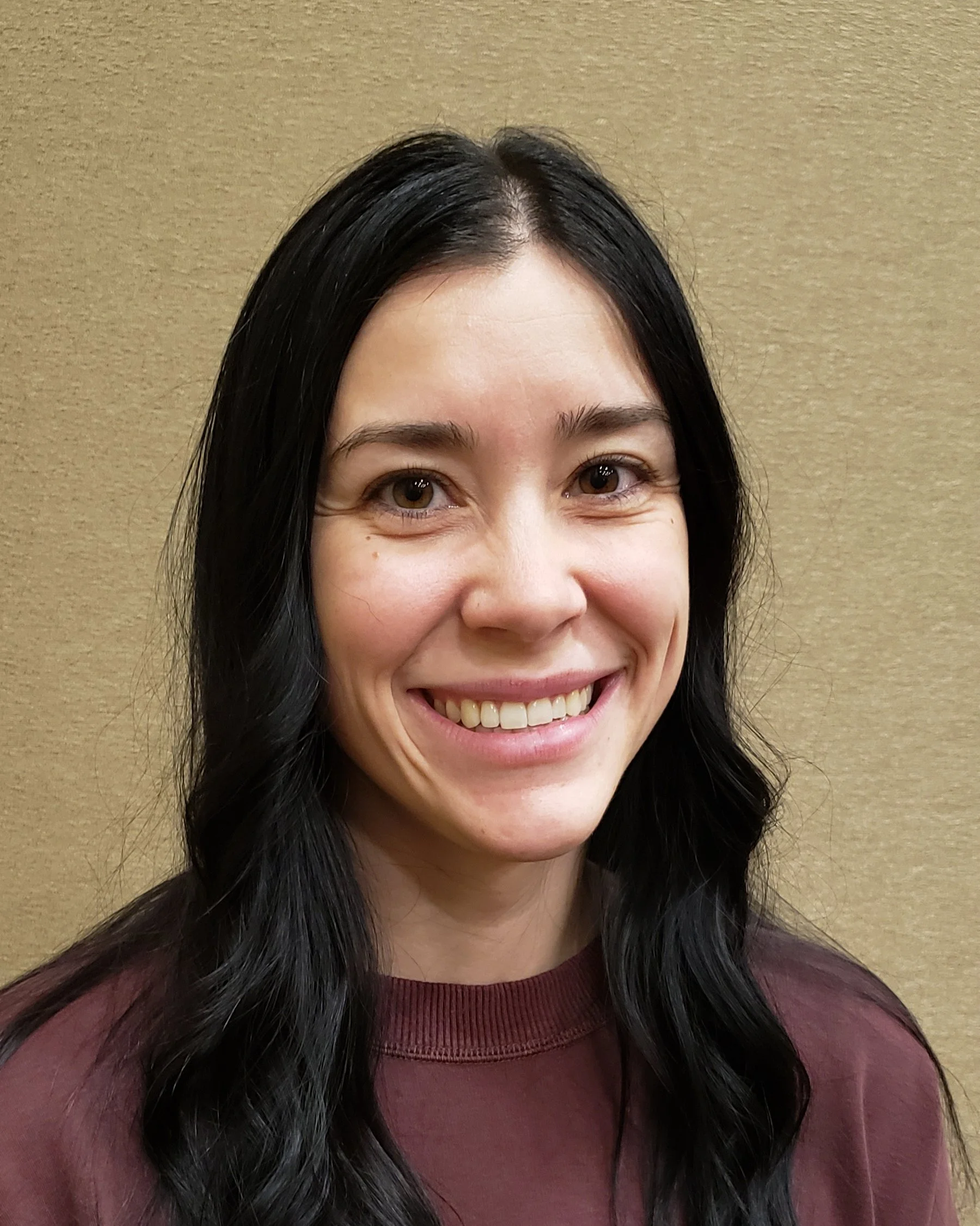 A woman with long black hair and brown eyes, smiling with teeth visible, wearing a maroon shirt, standing against a beige textured background.