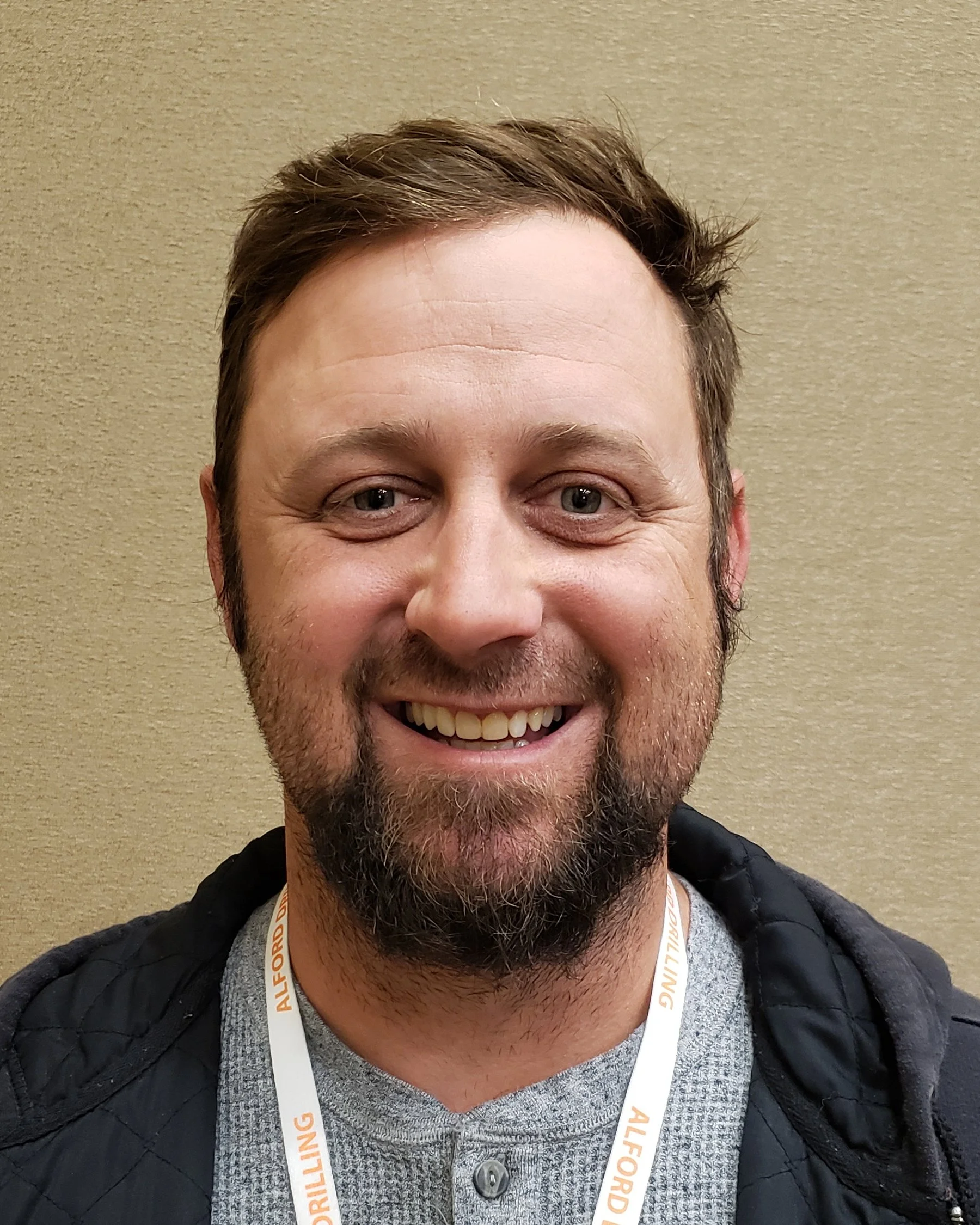 A smiling man with a beard and short brown hair, wearing a gray shirt and a black jacket, standing against a beige wall.