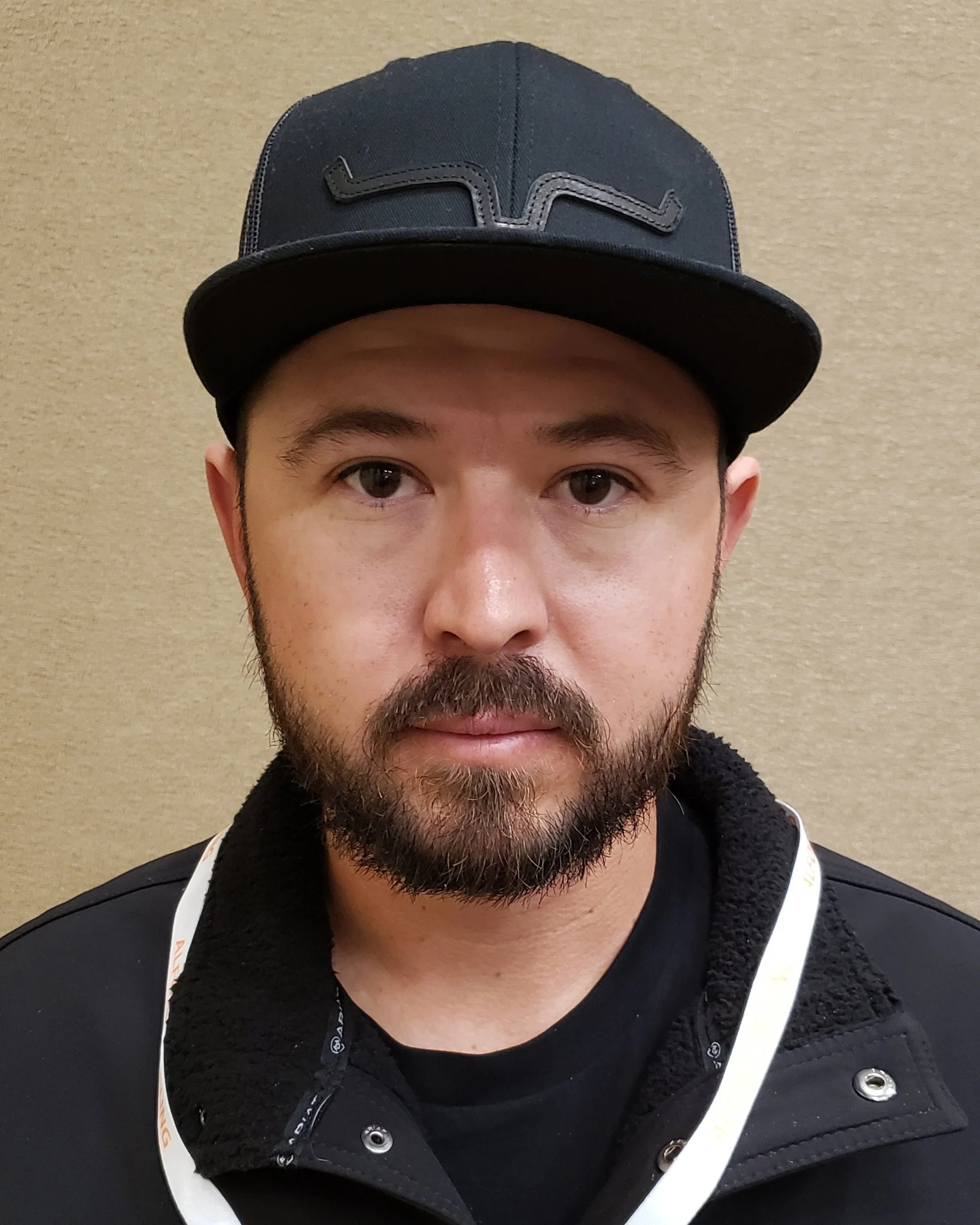 A man with a beard wearing a black baseball cap and black jacket in front of a beige wall.