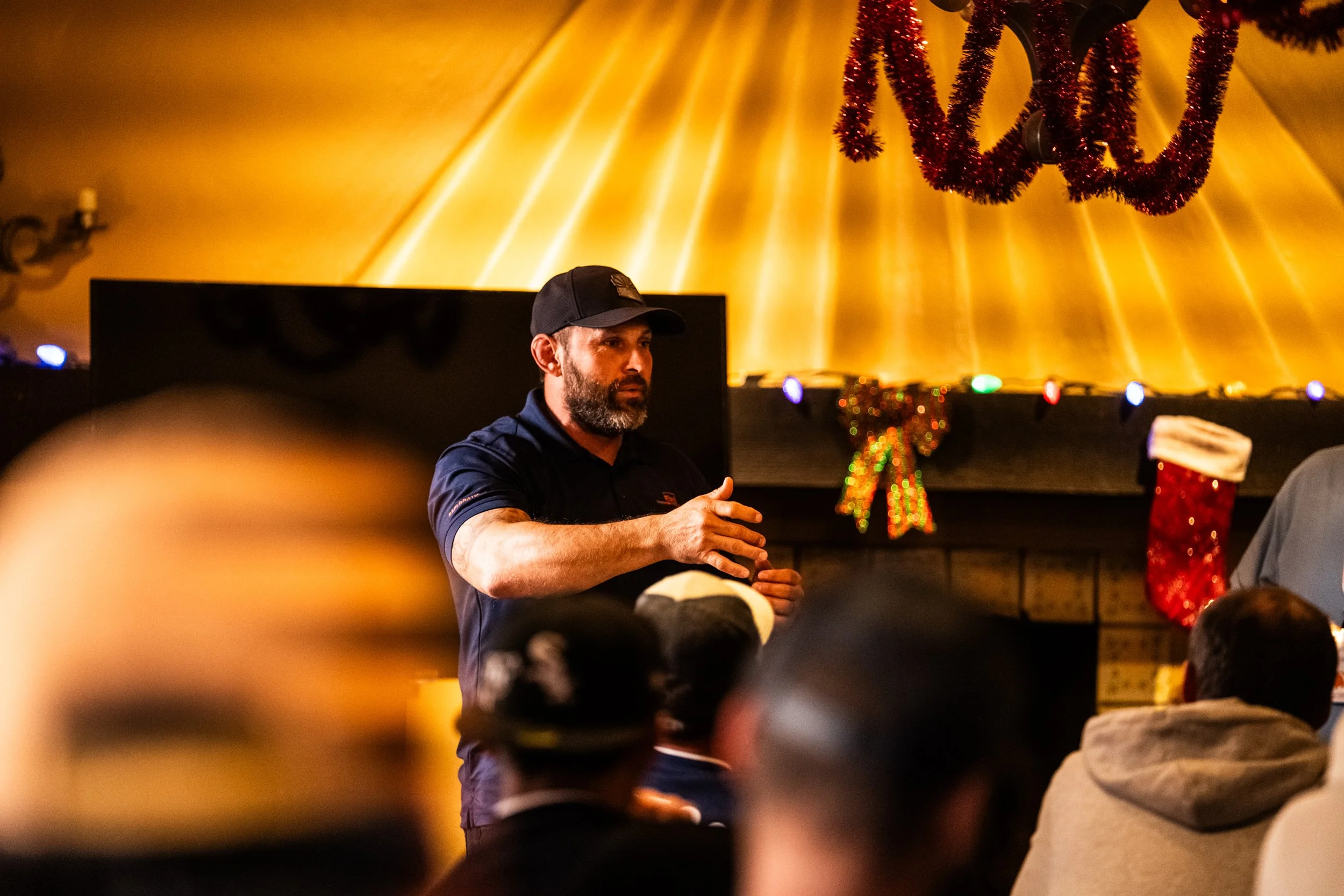 A man with a beard wearing a black cap and navy shirt giving a speech or presentation in a warmly lit room decorated for Christmas, with a fireplace, stockings, and Christmas ornaments.