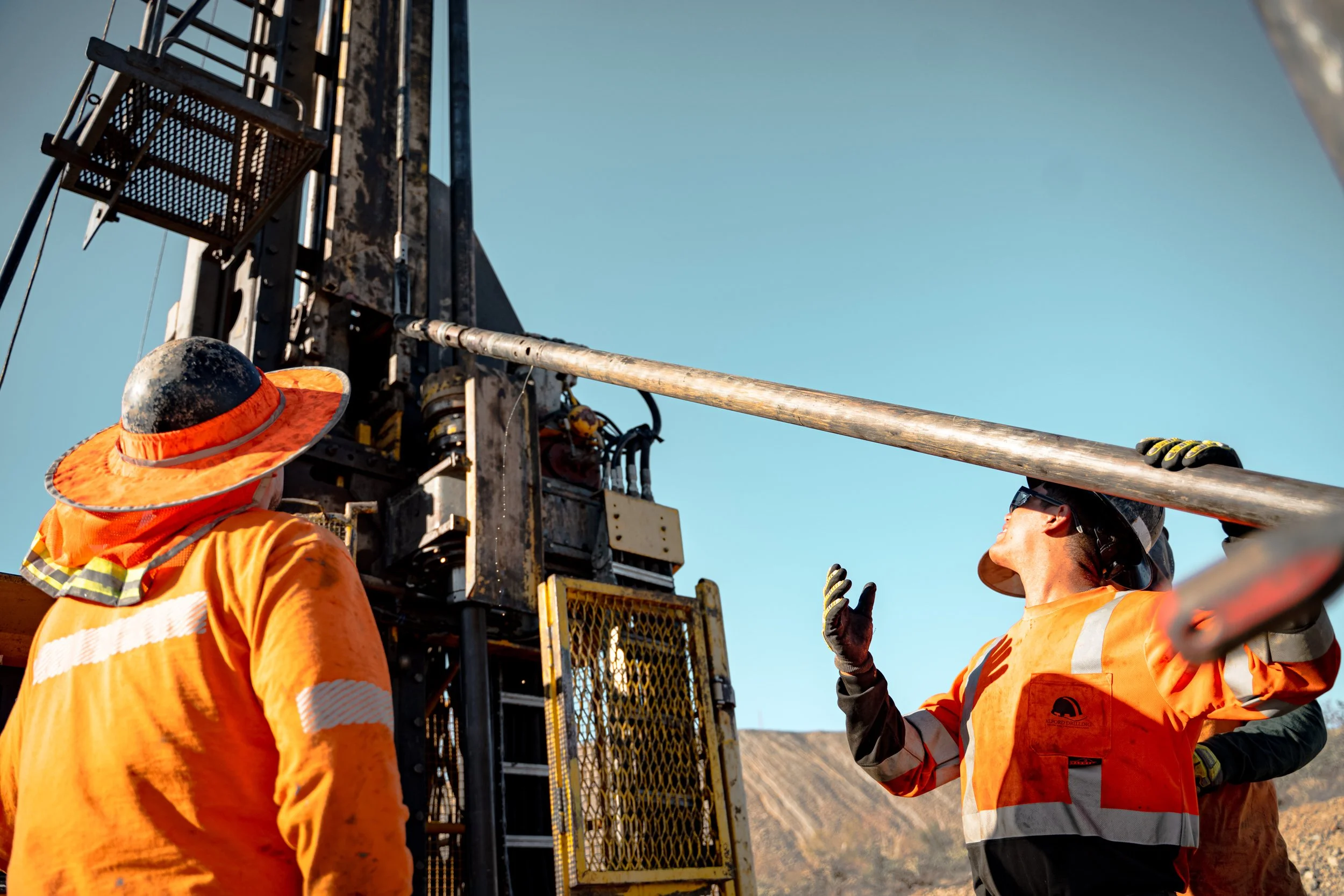 Two construction workers in orange safety vests and hats, working on a large piece of industrial equipment outdoors under a clear blue sky.