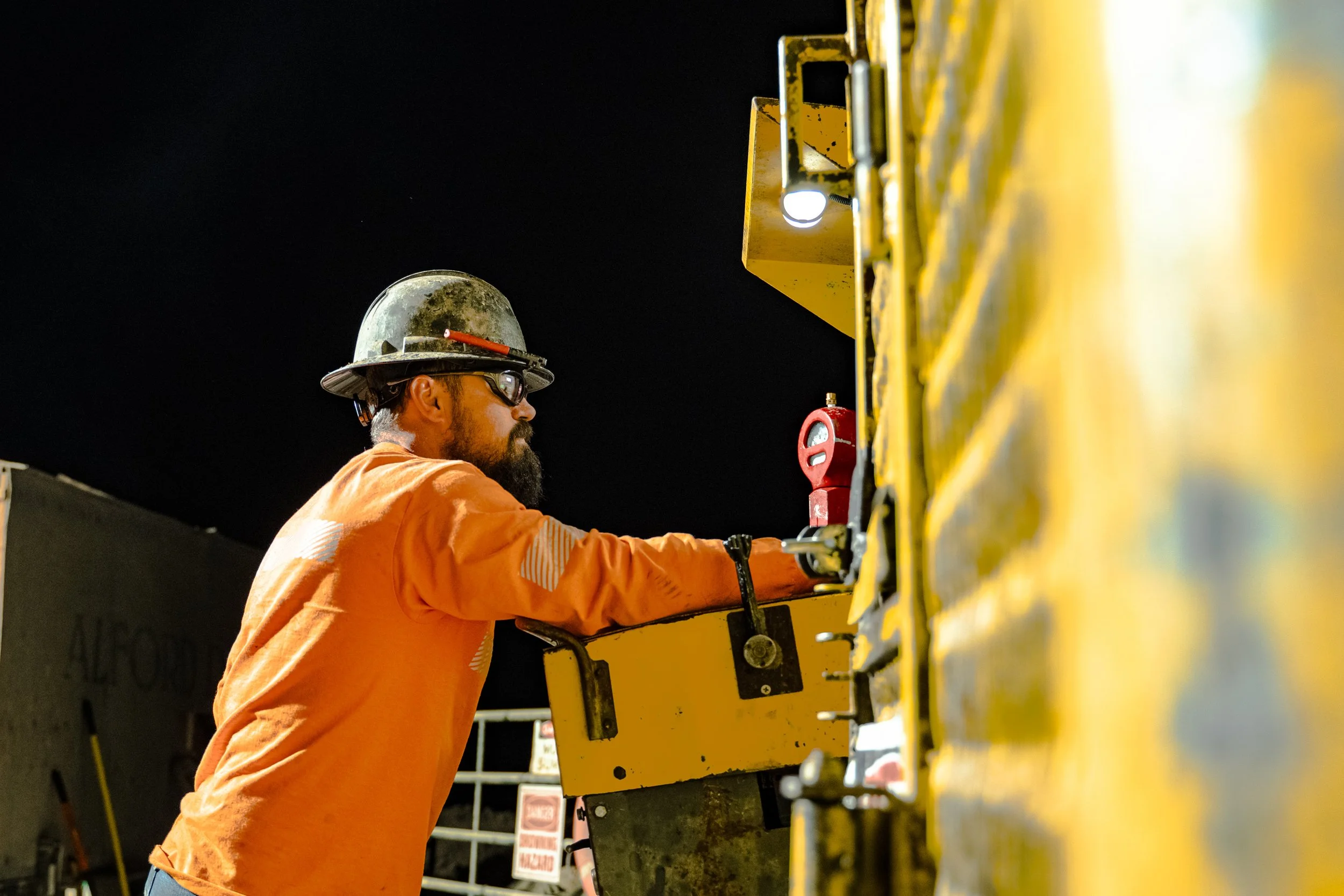 Worker wearing safety helmet and glasses, working at night on a yellow industrial machine.