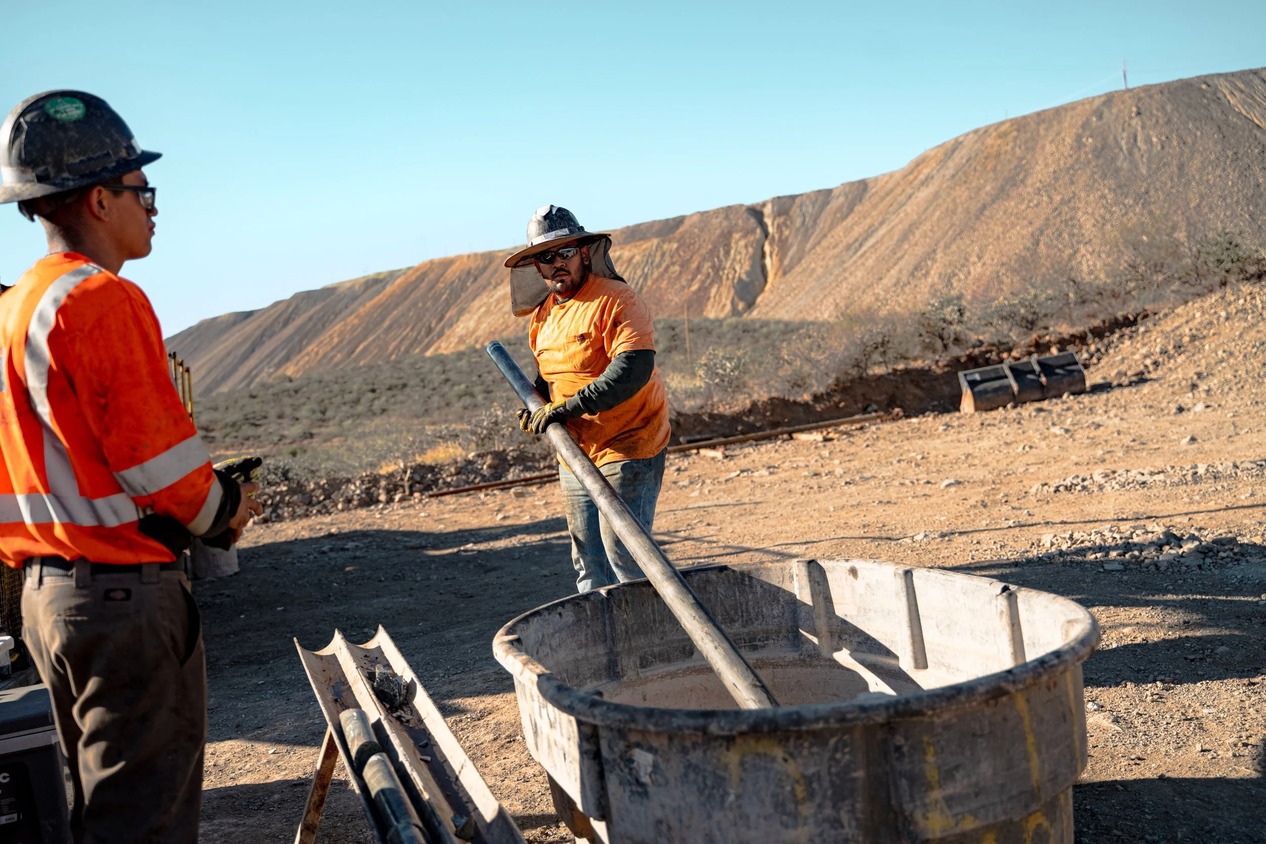 Two construction workers in safety gear working outdoors on a dirt site with mountains in the background.
