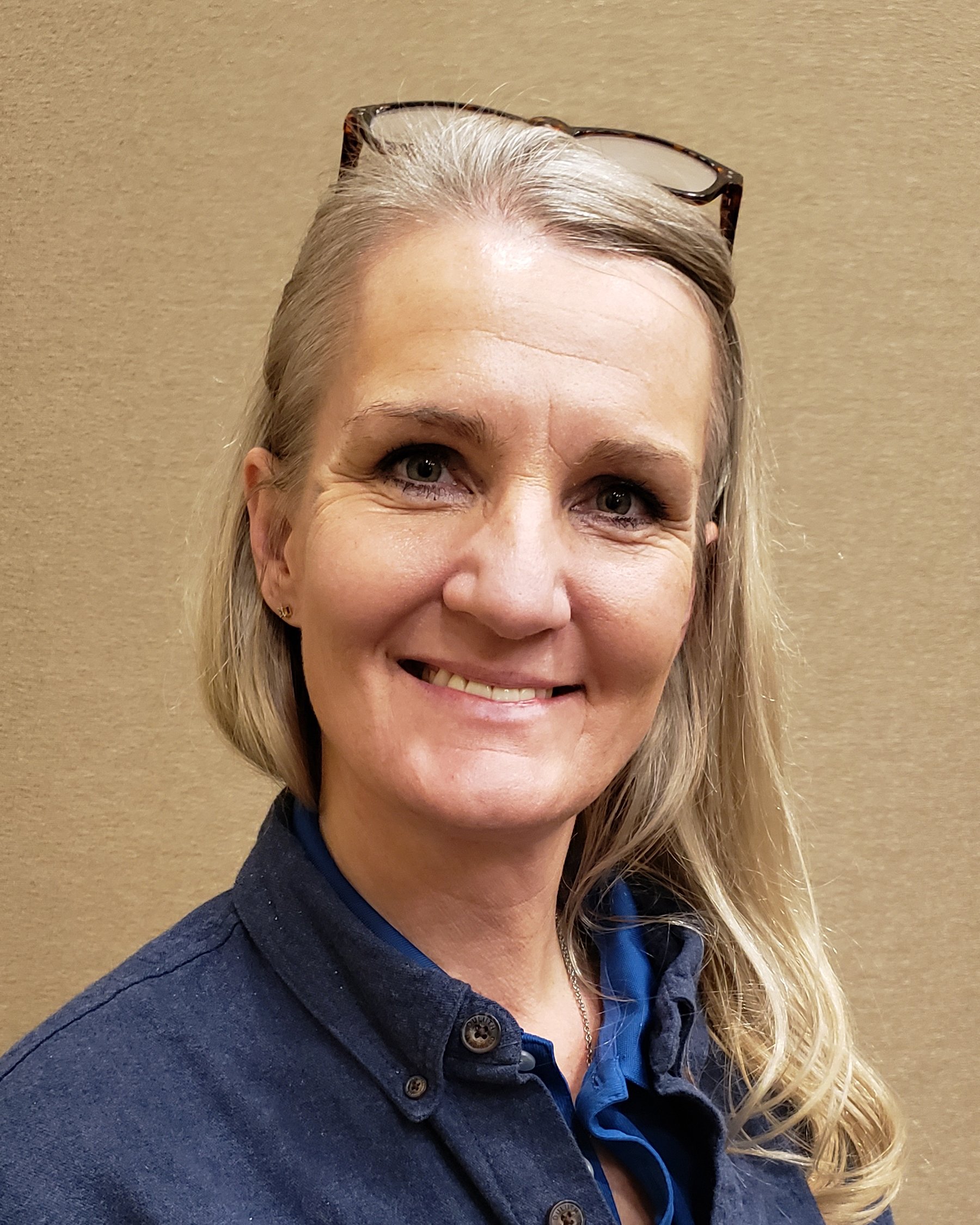 A woman with blonde hair wearing glasses on her head, smiling, in front of a plain beige wall.