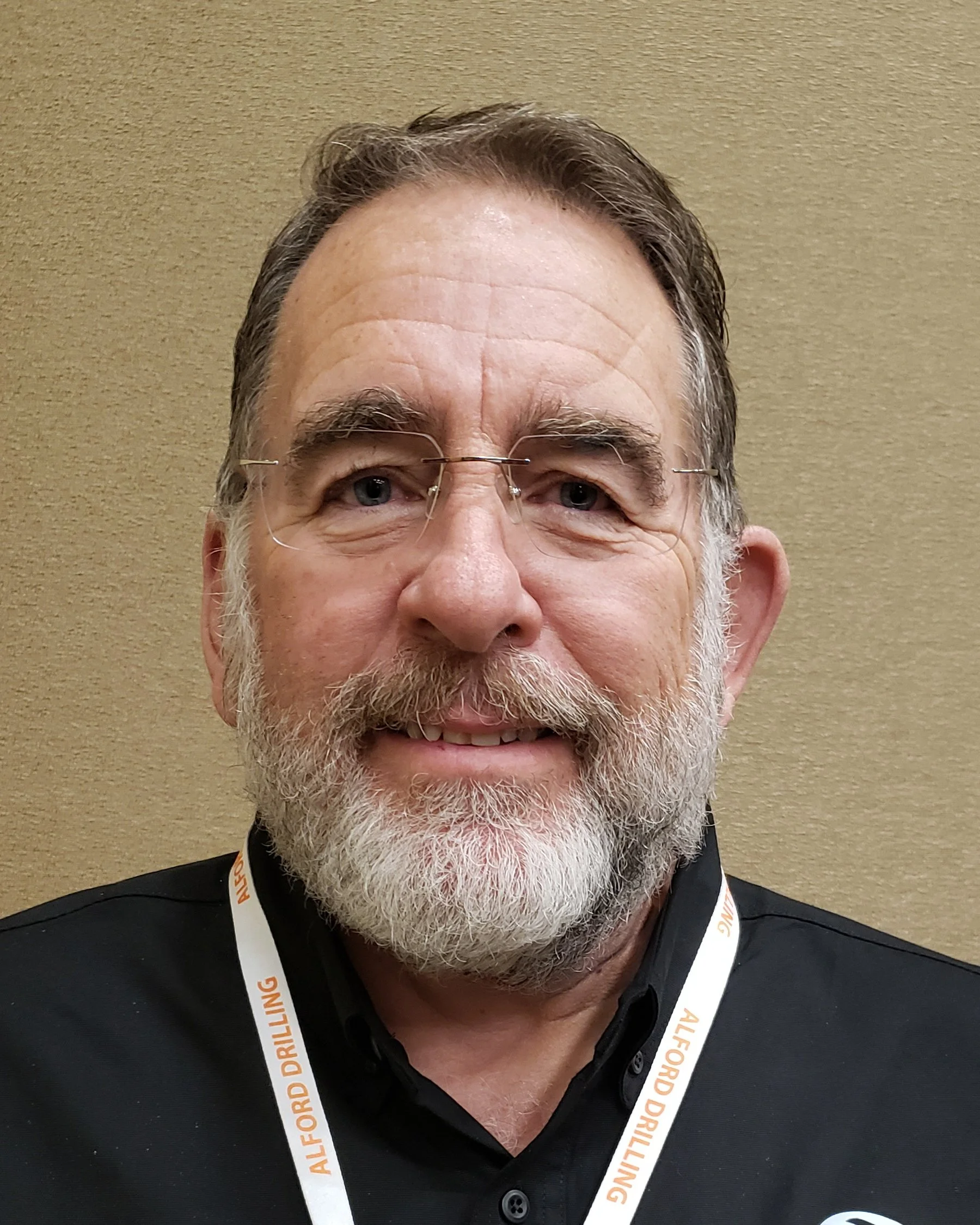 A smiling middle-aged man with gray hair and beard, wearing glasses and a black shirt, standing against a beige wall, with a white lanyard that reads 'ALFORD DRILLING' around his neck.
