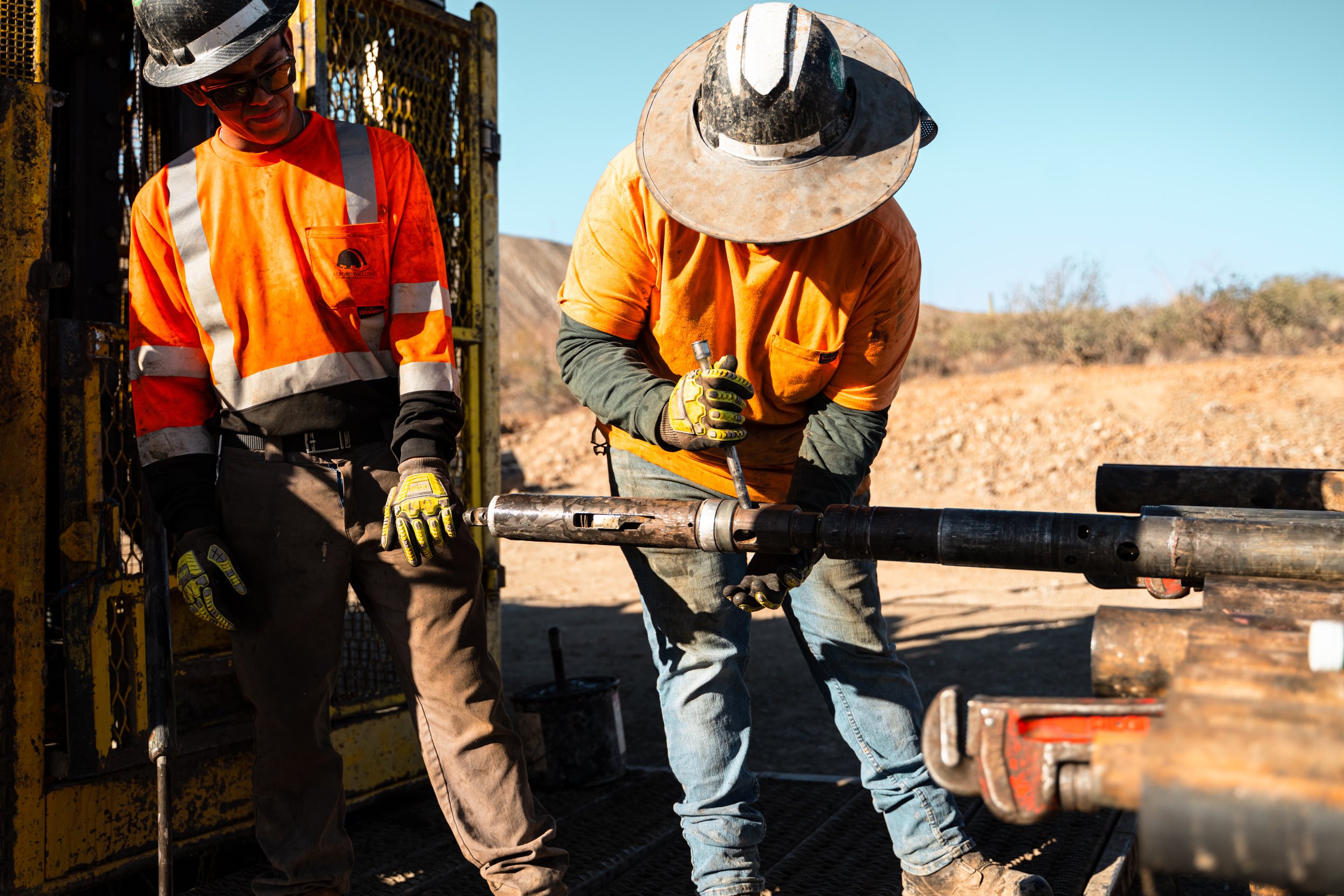 Two construction workers in orange safety vests and hats working outdoors with machinery and pipes, with a clear desert sky and dry landscape in the background.